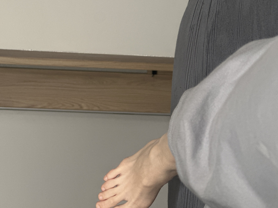 The scene is an indoor environment, identified as being in Yekaterinburg, Russia. The image displays a close-up, low-angle perspective of a room. In the foreground, a bare human left foot with fair skin is visible, extending from or resting on light grey fabric, which appears to be clothing or bedding. This fabric lies over a dark grey, horizontally ridged or pleated surface, consistent with bedding. The upper-left background features a plain light grey wall. To its right, a vertical wooden panel with a light brown, distinct grain pattern is visible, bordered by a thin metallic strip on its left edge, suggesting a cabinet door or wardrobe. A section of a plain off-white wall is visible to the right of the wooden panel, with a small dark detail, possibly a handle or gap, where the panel meets the wall. The subject's position suggests they are lying down.