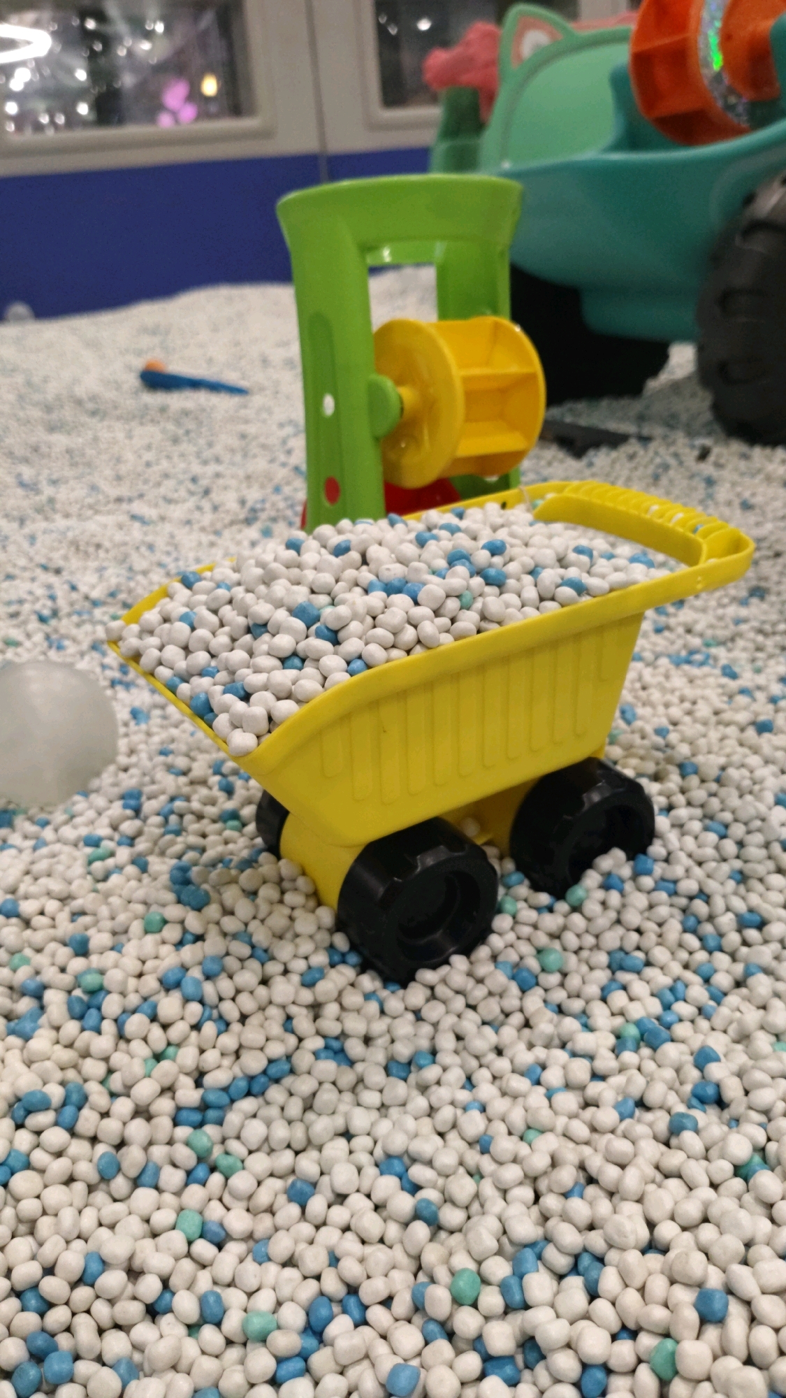 A close-up, eye-level shot shows a yellow toy dump truck filled with white and blue small round objects, possibly beads or candies, placed in a sandbox. The dump truck has black wheels and sits on a surface covered in the same white and blue objects. In the background, a green toy crane with a yellow rotating wheel is visible. Further in the background, a turquoise toy vehicle with large black wheels is partially seen. The environment appears to be an indoor play area. There are no people visible in the image. The lighting suggests an indoor setting with artificial lights, possibly with some reflections in the background.