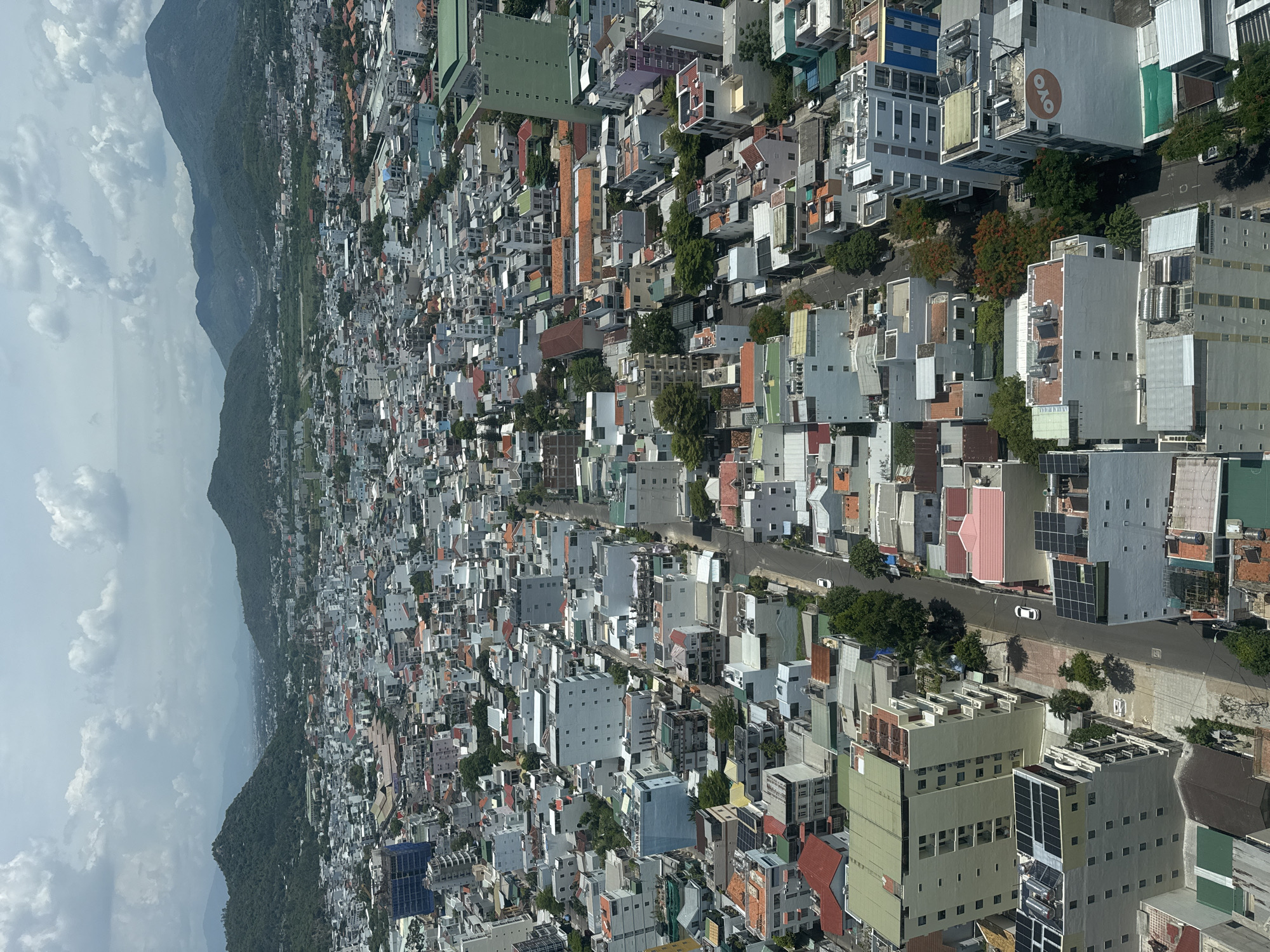 An aerial, high-angle, full-shot captures a dense urban landscape in Bac Nha Trang Ward, Vietnam. The scene is dominated by a vast expanse of buildings, ranging in height and architectural style, packed closely together. Buildings are a mix of white, grey, and pastel colors, with some featuring red-tiled roofs and green accents. Solar panels are visible on the roofs of several buildings, indicating a focus on sustainable energy. Trees are interspersed throughout the city, providing pockets of greenery. Roads weave through the city, with a few white cars visible, suggesting ongoing activity. In the background, rolling green hills and mountains rise against a cloudy sky, framing the urban sprawl. The overall impression is of a bustling, developing city nestled within a scenic natural environment.