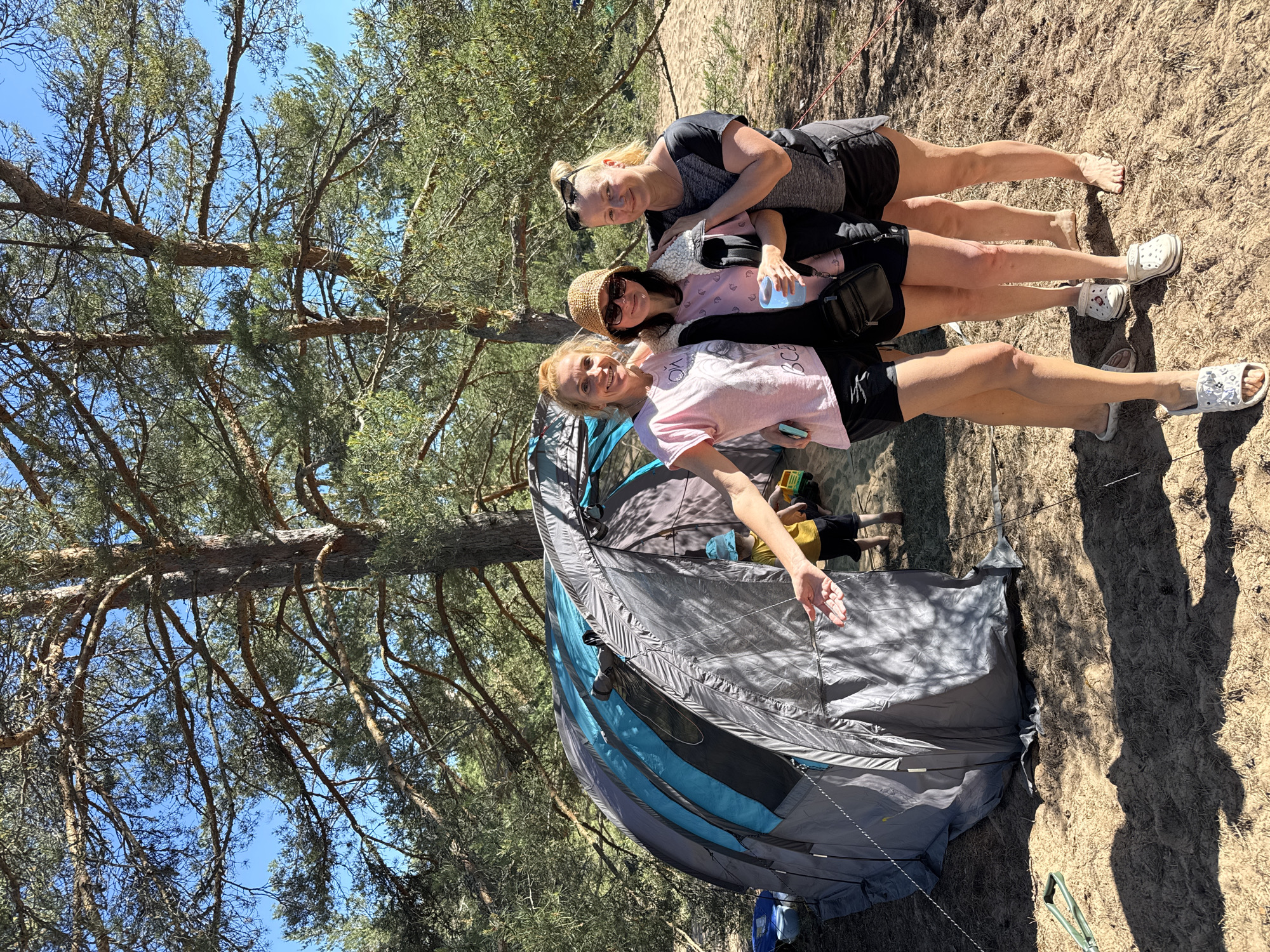 Three women stand in front of a grey and blue tent on a sandy area surrounded by trees. The woman on the left wears a pink t-shirt and black shorts, her left arm extended towards the camera. The middle woman wears a pink t-shirt, black jacket, and a straw hat, holding a blue cup. The woman on the right wears a black t-shirt and black shorts, with sunglasses on her head. A young child in a blue hat and yellow shirt is partially visible inside the tent. The environment appears to be a campsite in a wooded area with sandy ground. The sky is blue and sunny.
