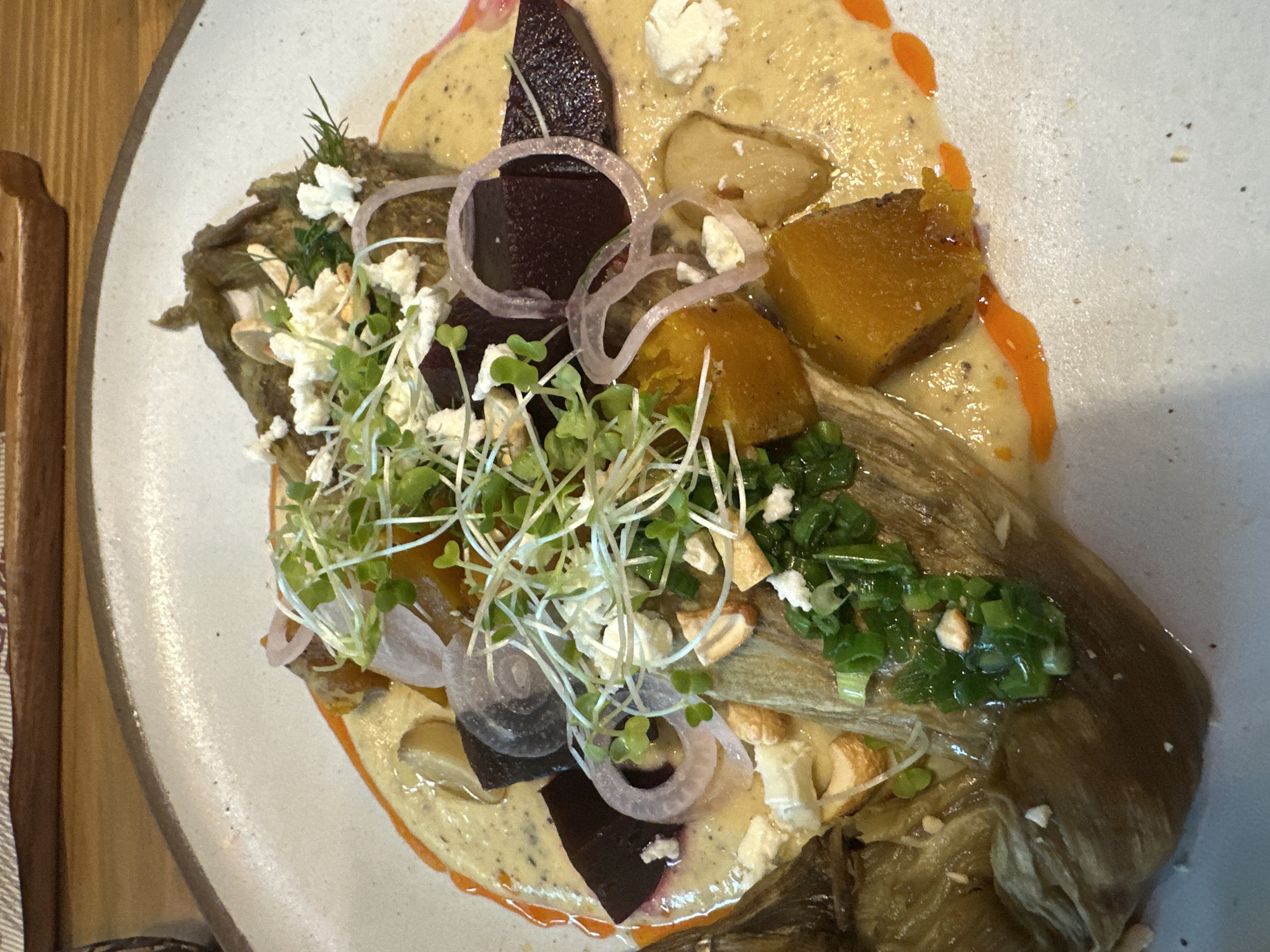 The image presents a close-up, high-angle view of a meticulously plated dish served on a round, off-white ceramic plate with a speckled texture. The dish appears to be a gourmet vegetable preparation, possibly an appetizer or a light main course.
A creamy, pale yellow sauce or purée, speckled with dark spices, forms the base, artfully spread across the plate. Bright orange streaks or drizzles of a complementary sauce or oil are visible, particularly along the edges of the main sauce, adding a vibrant accent.
Layered on this base are several roasted vegetables. Large, elongated pieces of a soft, brownish vegetable, likely roasted eggplant, anchor the composition. Scattered across the dish are rich, dark purple chunks of roasted beets and bright, golden-orange cubes, which appear to be roasted squash or sweet potato. Delicate, translucent rings of thinly sliced red onion are strategically placed, providing a visual and textural contrast.
The dish is generously garnished with fresh elements. A prominent cluster of slender, light green sprouts or microgreens is centered, adding height and a fresh appeal. Chopped green onions or chives are sprinkled in various sections. Further garnishes include small crumbles of white cheese, possibly feta or goat cheese, and small, light-colored nut pieces, such as slivered almonds or cashews, contributing to texture. Tiny sprigs of green herbs, possibly dill, are also visible.
The overall presentation suggests a thoughtful and artistic approach to cooking. The background reveals a wooden surface to the left, likely a table or serving board. No people are visible in the frame, and there is no discernible text. The lighting is bright and even, highlighting the dish's vibrant colors and textures.