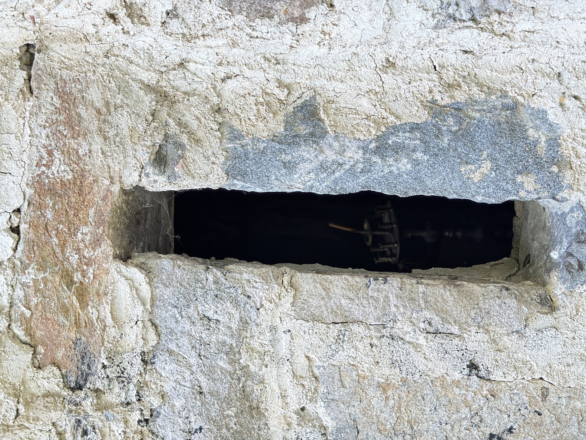 A stone wall in Govedartsi, Bulgaria, contains a rectangular opening. Within the opening, a dark space is visible, with a metal gear and rod partially visible. The stone wall is textured, featuring variations in color from gray to white with some hints of brown. Cracks and crevices are evident on the wall's surface.