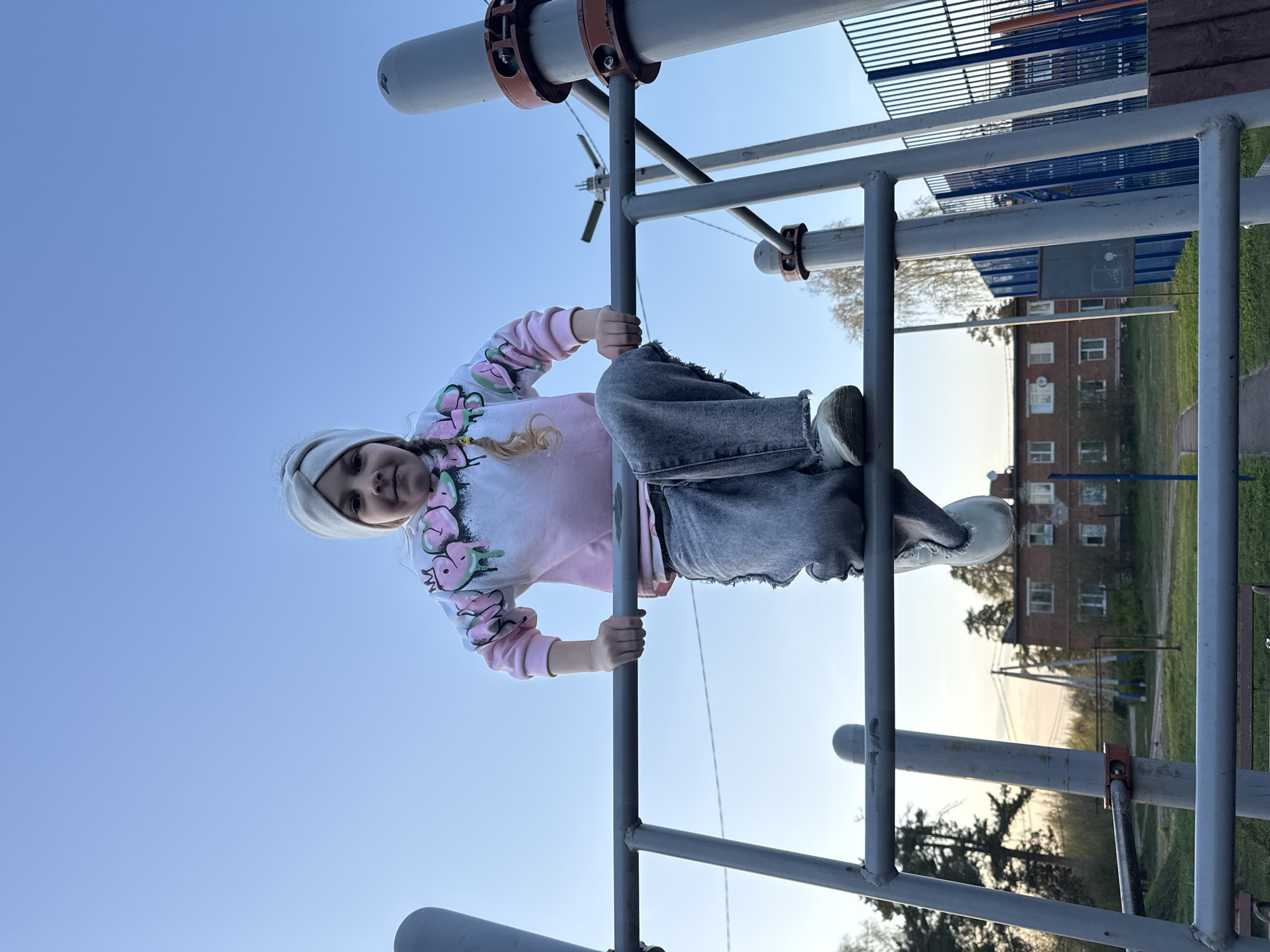 A young girl is hanging upside down from a metal jungle gym structure, holding onto the bars with her hands. She is wearing a light-colored beanie, a pink and white tie-dye style sweatshirt with watermelon designs, grey jeans, and white shoes. The background reveals a cityscape with buildings of a brick hue and blue structures, suggesting an urban location. The photo was taken outdoors, under a bright sky. Based on the location metadata, the image was taken in Раздолье, Russia.