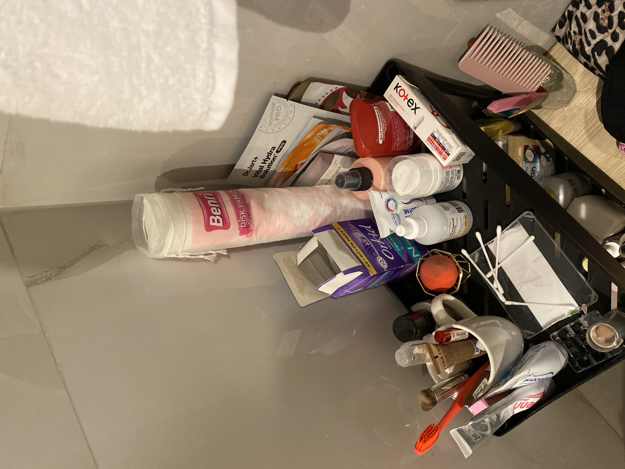 This image depicts a black plastic multi-tiered shelf unit filled with various personal care and cosmetic items. The shelf is positioned in what appears to be a bathroom or vanity area, with a light-colored tiled wall and a white fuzzy towel hanging on the right.
On the top shelf, a large cylindrical package of "Benri Disk Pamuk Round Cotton" is prominent. To its right, a folded "Dr.Jart+ Vital Hydra Solution PRO" mask packet is visible, along with other mask packets. Below these, a purple box labeled "Orkid" is partially open, containing what looks like feminine hygiene products. A small red, spherical object is placed on a gold geometric stand. There's also a light pink spray bottle and a white jar with a white lid.
The middle shelf holds a white mug containing several items, including an orange toothbrush, makeup brushes, a tube of "Flormar," and another red tube. A clear plastic bottle with a white cap is on the far left, and a black bottle with a black cap is to the right of the mug. A red container is also on this shelf. A white box with red and blue lettering, labeled "Kotex," is positioned on the right side of this shelf.
The bottom shelf contains a clear plastic tray with cotton swabs, a black plastic tray with what appears to be a makeup compact or similar item, and several bottles and packages. A pink comb is standing in a clear glass on the right side of the bottom shelf, next to other small items. A patterned fabric, possibly a leopard print, is visible on the lower right corner.
The overall environment suggests a personal storage space for beauty and hygiene products. The presence of specific brands like "Benri," "Dr.Jart+," "Orkid," and "Kotex" indicates common consumer products. The scene is static, with no visible human interaction.
While the image context strongly suggests a private residential or hotel bathroom setting, it is not possible to determine its specific location within the city of Aksu, Turkey, based solely on the visual information provided.