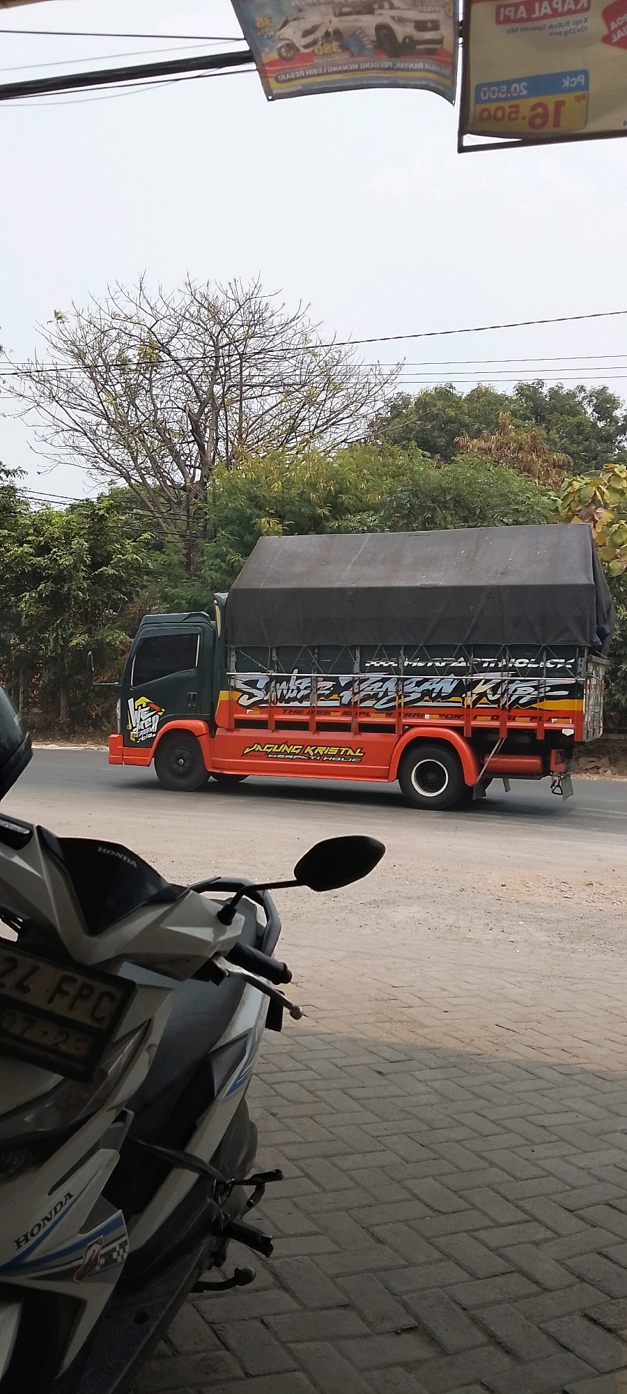 In Bekasi, Indonesia, a dark green and orange truck is parked on a paved road, its cargo area covered by a black tarp. The truck has text on its side that reads "WEAKEN KARTEL" and "JAGUNG KRISTAL HERBATI HOLIC" in white and red lettering. In the foreground, a white and grey Honda motorcycle is parked, with its license plate partially visible, reading "24 FPC". The background features trees and a cloudy sky.