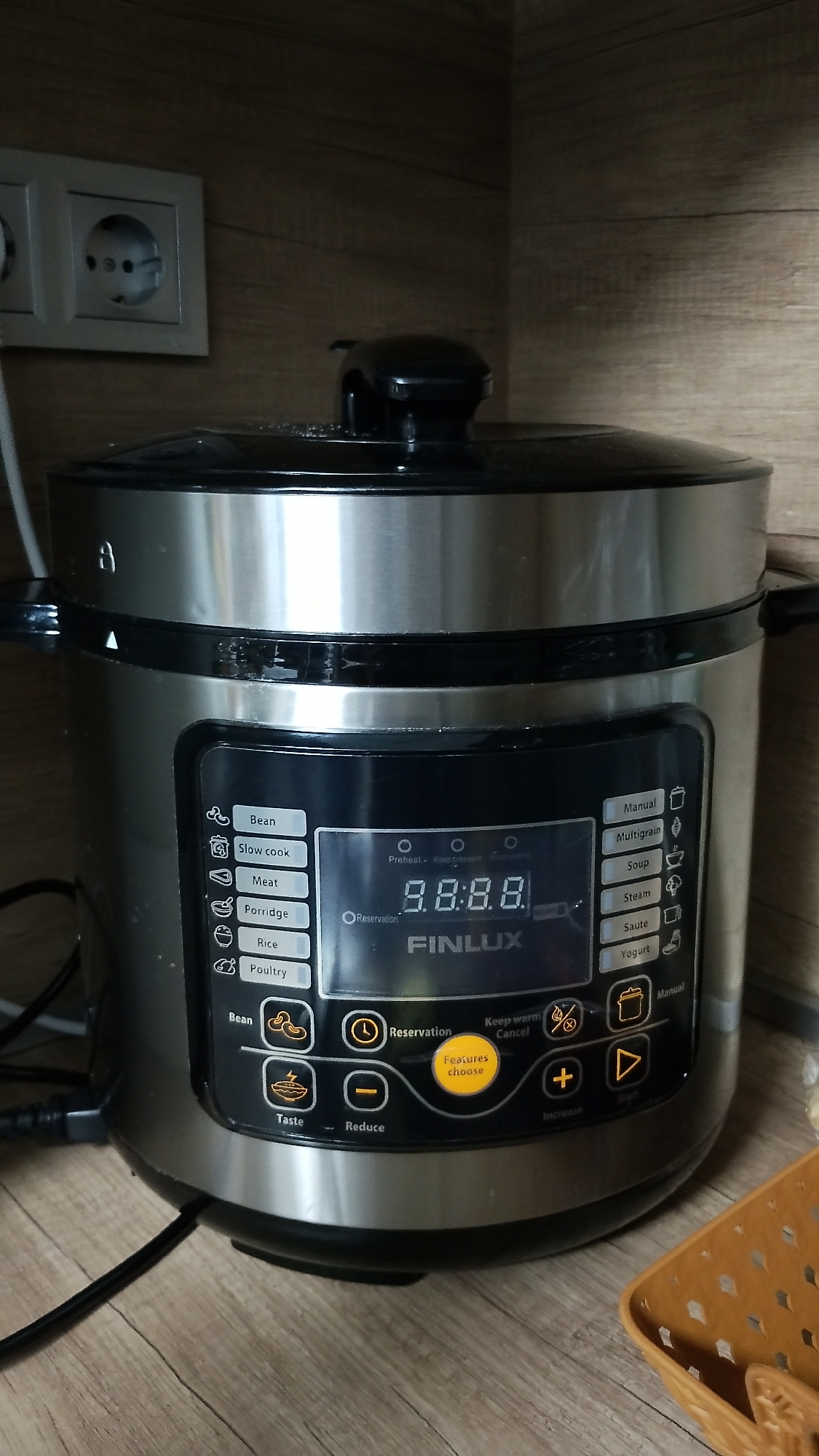 The image shows a silver and black multi-cooker appliance on a wooden surface, possibly a countertop. The appliance has a digital display showing "8.8.8.8" and text labels for various cooking functions like "Bean," "Slow cook," "Meat," "Porridge," "Rice," "Poultry," "Manual," "Multigrain," "Soup," "Steam," "Saute," and "Yogurt." There are also buttons labeled "Taste," "Reduce," "Increase," and "Start." The appliance is plugged in, with a black cord visible. In the background, there is a wooden wall with horizontal planking and a light-colored electrical outlet with two sockets. The overall scene is an indoor domestic setting. The location is not determinable from the image content.