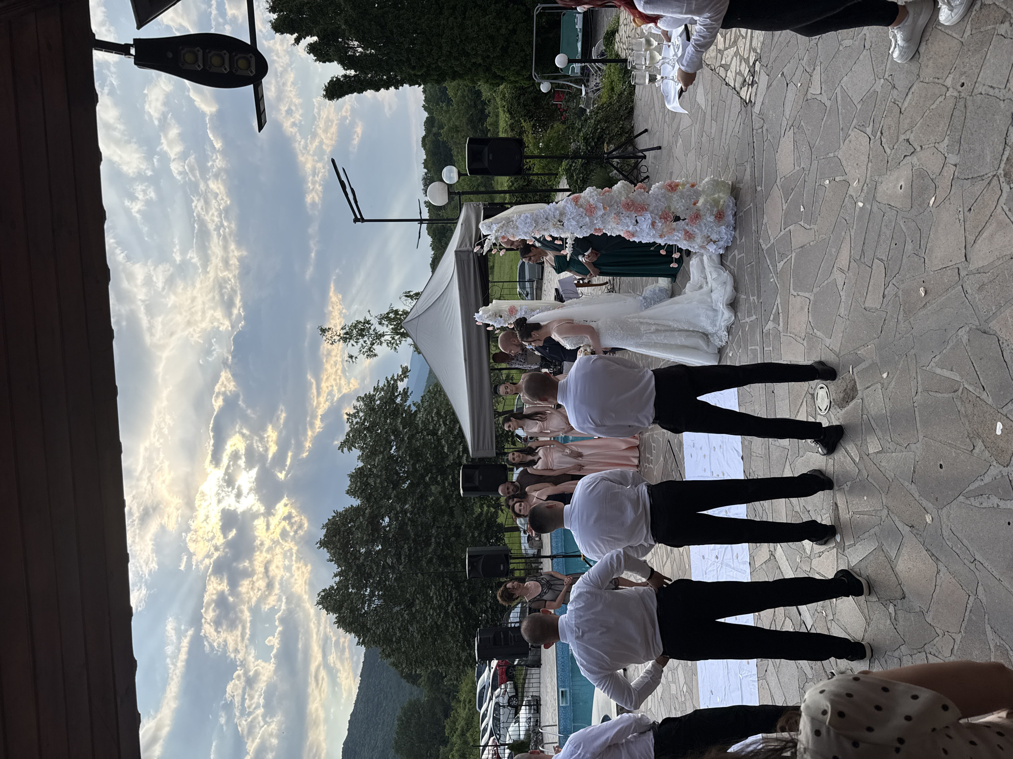 An elevated, downward-angled outdoor photograph captures a wedding ceremony or event in Debrashtitsa, Bulgaria, under a partly cloudy sky with visible sun rays. The scene is observed from beneath a dark wooden overhead structure.
In the immediate foreground, four men in white long-sleeved shirts and black trousers stand with their backs to the viewer, observing the event. To the far left, a person partially visible wears a white shirt with black polka dots. To the far right, another person, also with their back to the viewer, wears a white shirt, black trousers, and white sneakers, holding a white rectangular tray with four champagne flutes.
The main event unfolds on a stone-paved patio area. A white fabric runner is laid on the ground, leading to the central ceremony area, which is flanked by two tall, white, and pink floral decorative columns. A bride in a white gown and veil stands at the end of the runner, near a partially visible groom. Adjacent to them, a woman in a dark green dress, appearing pregnant, is visible. Several women in varying shades of pink and peach dresses (bridesmaids) are lined up to the left of the central figures. Another woman in a dark, patterned dress stands facing the group, possibly officiating.
Two large black loudspeakers on stands are positioned on either side of the ceremony area. A white tent provides cover to the right. In the mid-ground, a blue swimming pool is visible on the left, behind the observers. Beyond the pool and ceremony area, a parking lot contains multiple cars of various models and colors. The background features lush green foliage, tall trees, and rolling mountains under the cloudy sky. Architectural elements include a modern dark-colored solar-powered street light attached to an unseen structure on the upper right, and a standard street light further back. White flower petals are scattered across the stone patio. A swing set with a light blue seat and two spherical white outdoor lights are partially visible in the far right background, nestled among trees.