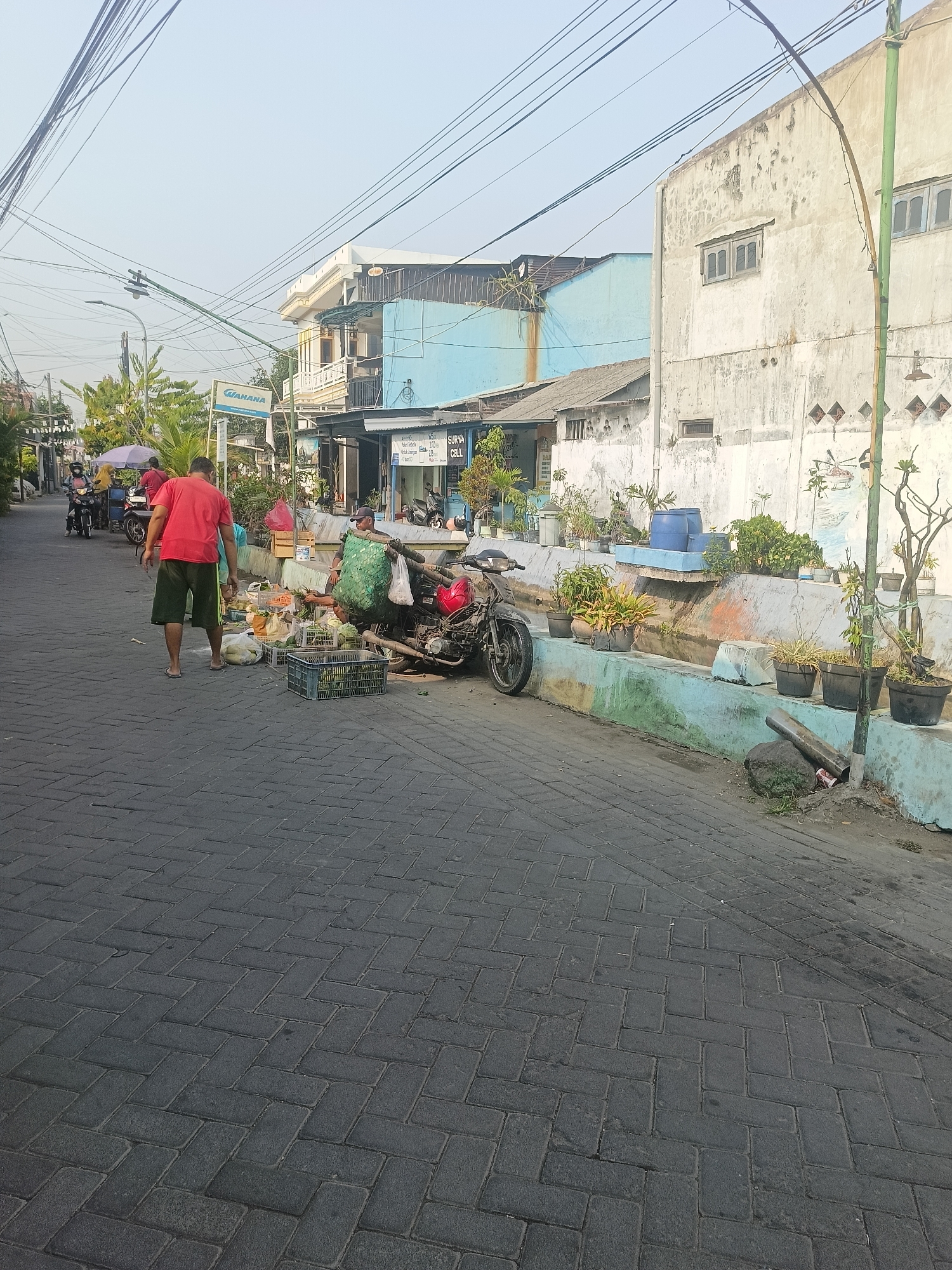 The image captures a street scene in Gedangan, Indonesia, likely in the morning due to the soft lighting. The street is paved with dark gray interlocking brick pavers laid in a herringbone pattern. To the left of the image, a few people are seen on motorcycles, and one person is walking away from the camera, wearing a red shirt and green shorts.
In the foreground and middle ground, a street vendor is present, with a motorcycle parked near a collection of fruits and vegetables displayed in crates and bags. The vendor, wearing a green sack over their shoulders, is interacting with a customer or arranging the produce.
The environment includes buildings lining the street. On the right, a weathered, light-colored building with visible signs of age and perhaps some graffiti or murals is prominent. To the left of this building, a lower, light blue building is visible, followed by a multi-story structure with balconies and windows. Overhead, a complex network of electrical wires crisscrosses the sky, typical of urban or semi-urban Indonesian settings. Planters with various plants are placed along a low concrete wall separating the sidewalk from a shallow, dry ditch. The overall impression is of a bustling, everyday street in a residential or commercial area.