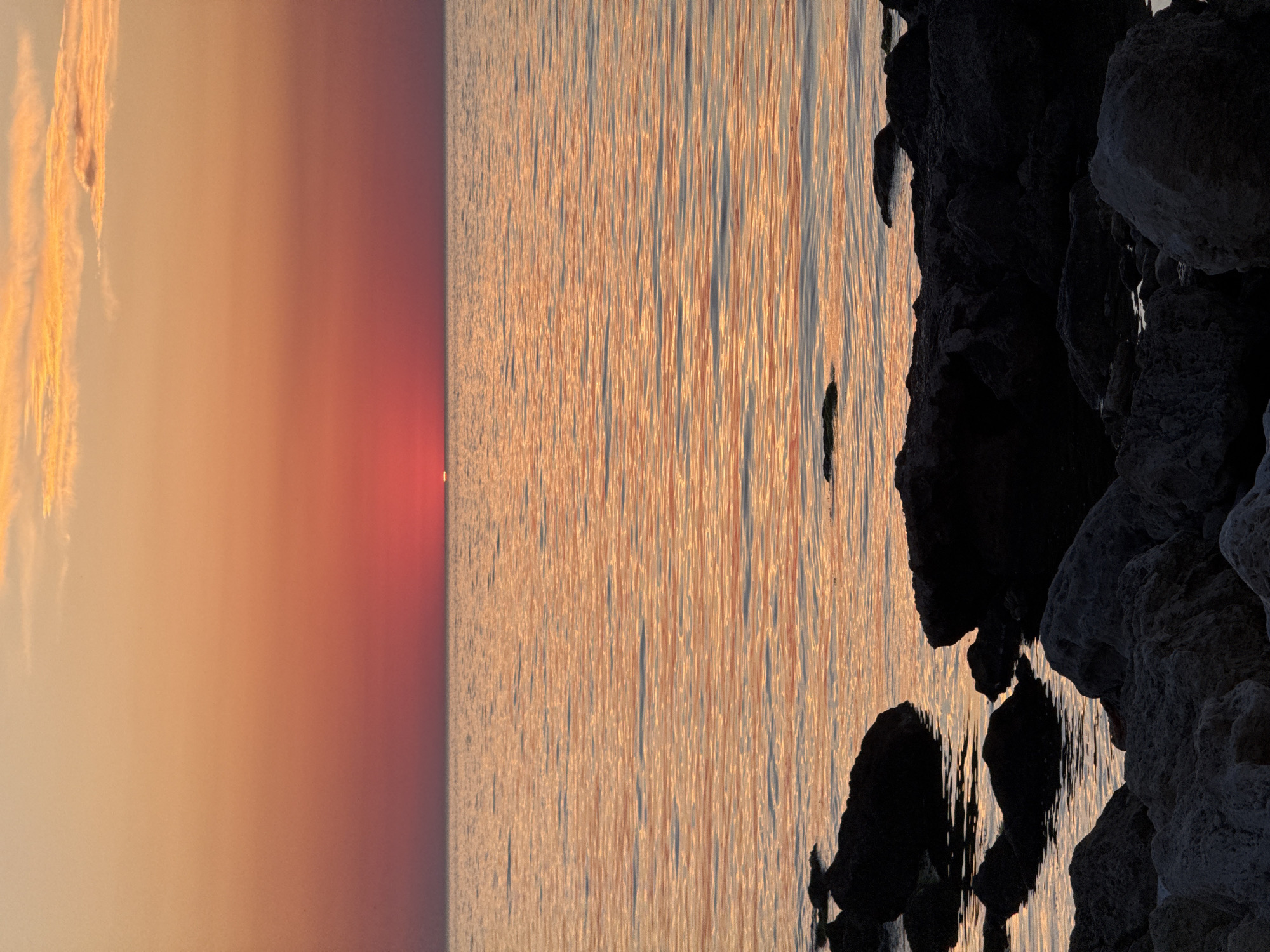 An overhead, close-up shot captures the reflection of an orange and red sunset on calm ocean water. The sun is visible as a small, bright orange disk on the horizon. In the foreground, dark, silhouetted rocks are partially submerged in the water, with more rocks and a rocky shoreline visible further back. The sky above transitions from a deep orange at the horizon to a lighter orange and then a pale yellow with wispy clouds near the top of the frame.
