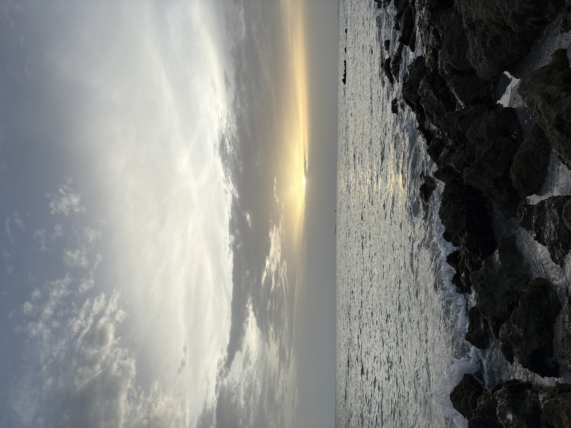 The image depicts a serene seascape at dusk, likely near Venice, Florida, United States of America, given the presence of a rocky shore and the characteristic calm waters of the Gulf of Mexico. The sky is a blend of soft grays and pale yellows, with the sun partially obscured by scattered clouds, casting a gentle, diffused light. The ocean stretches towards the horizon, its surface rippled by a light breeze, reflecting the muted hues of the sky. A rocky shoreline dominates the foreground, with dark, irregularly shaped stones leading into the water. Small waves crash softly against the rocks, creating a dynamic contrast with the stillness of the sea and sky. There are no people visible in the scene, and the overall atmosphere is one of peaceful solitude and natural beauty. The image is a still, observational capture of a natural landscape.
