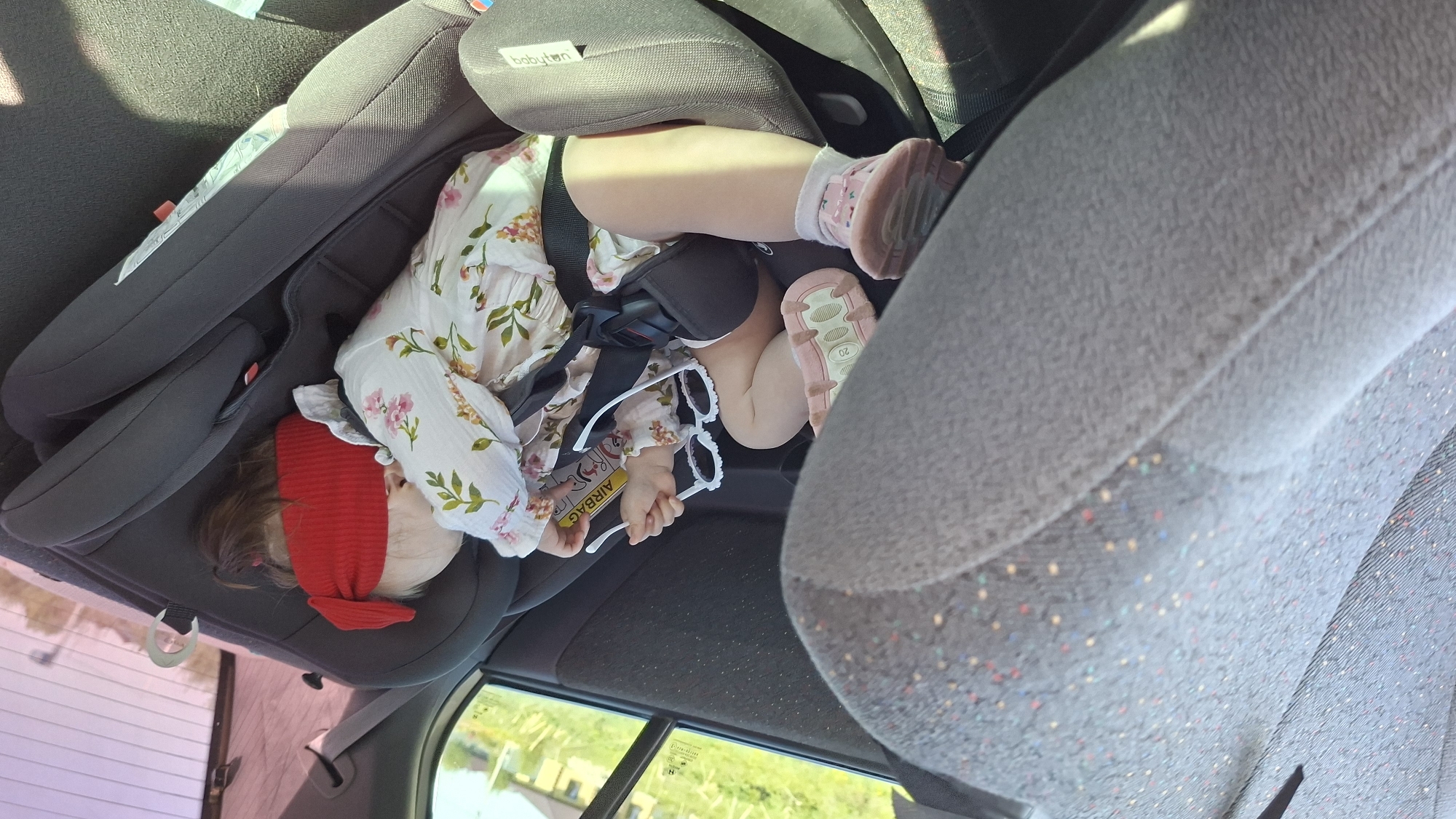 A baby is asleep in a Babyten brand car seat in what appears to be a car interior. The baby is wearing a floral dress, white socks, and pink shoes. A red headband is on the baby’s head. The baby is buckled into the car seat with a black harness. The baby holds a pair of white, round sunglasses in their hand, near a yellow tag with the word "Airbag" printed on it. An adjacent car seat, upholstered in gray fabric with multicolored speckles, is visible. Outdoor scenery, including green vegetation, is seen through the car window, but the location (Гаревая, Russia) is not visually verifiable from the image alone.