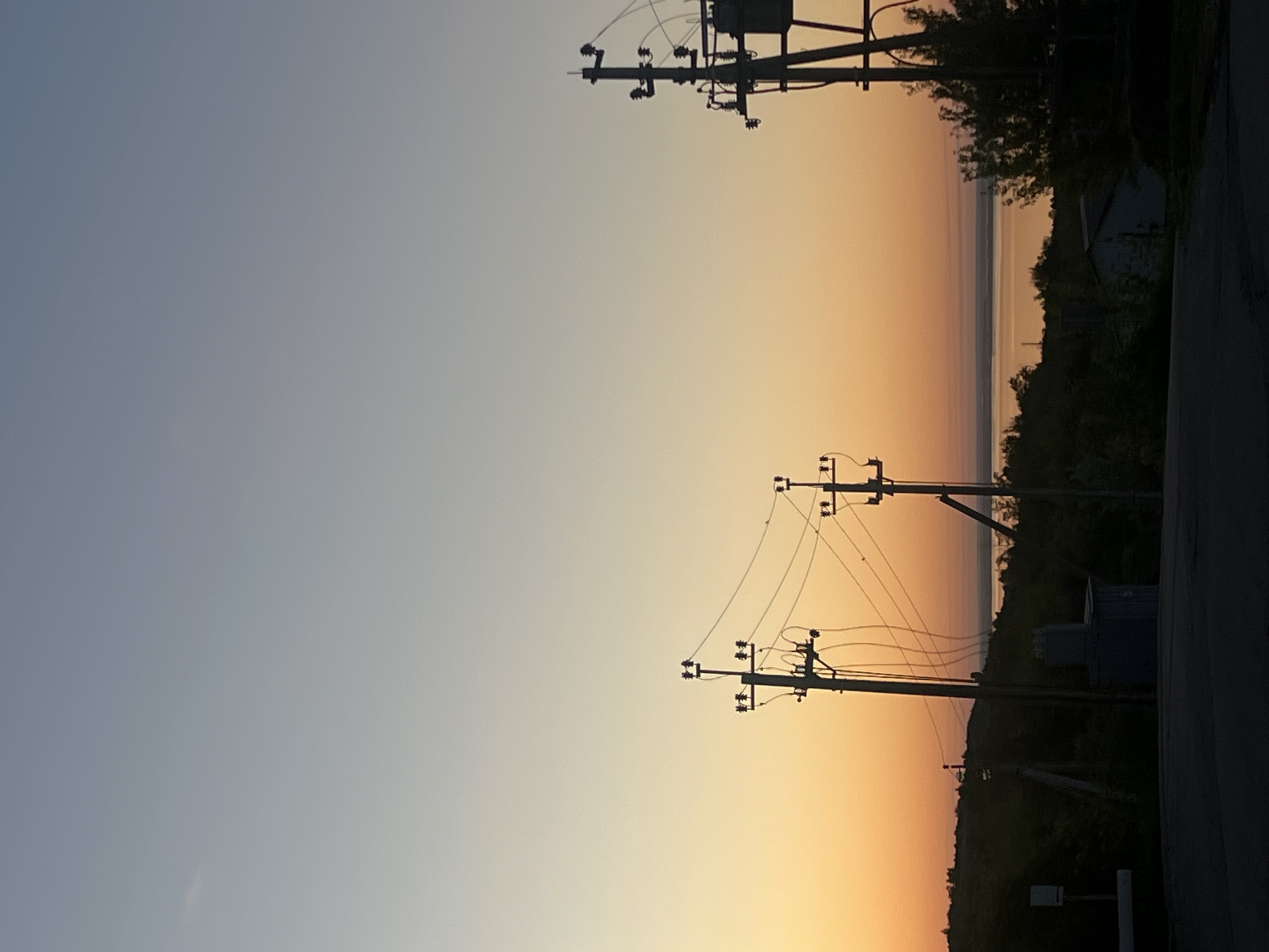 The image shows a sunset scene in Khvalynsk, Russia. The sky transitions from a pale blue at the top to a warm orange near the horizon. Two sets of utility poles and wires are visible against the sky. The horizon line shows the edge of a body of water, possibly the Volga River, with trees and some structures lining the shore. A road and some greenery are visible in the lower right corner of the frame. The general impression is of a quiet evening with the sun setting over the river landscape.