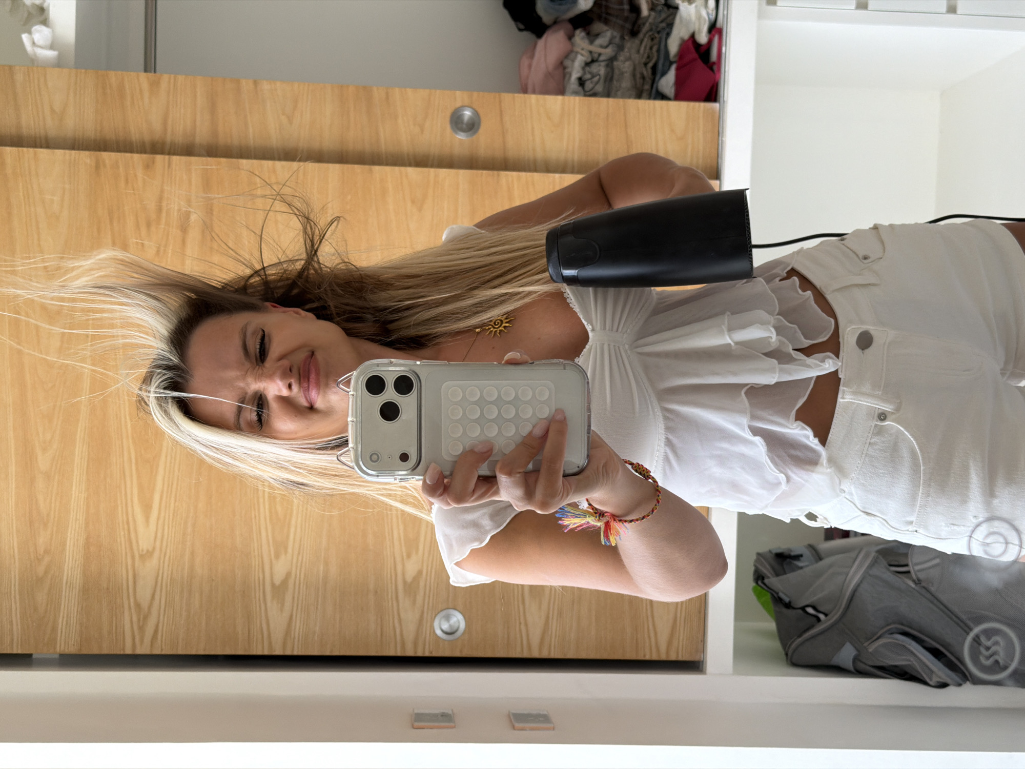 A young woman with blonde hair, wearing a white ruffled top and white shorts, is taking a mirror selfie in what appears to be a closet or dressing area. Her hair is being blown upward, creating a dynamic effect. She is holding a smartphone in her left hand, which is positioned to capture the reflection. In her right hand, she holds a black hairdryer, which is likely the cause of her hair's disarray. She is wearing a gold sun pendant necklace and a colorful braided bracelet on her left wrist. The background features wooden closet doors and shelves containing clothing. The presence of a grey baby carrier or backpack is visible on a shelf to the left. The overall impression is a candid and playful moment captured in a personal space.