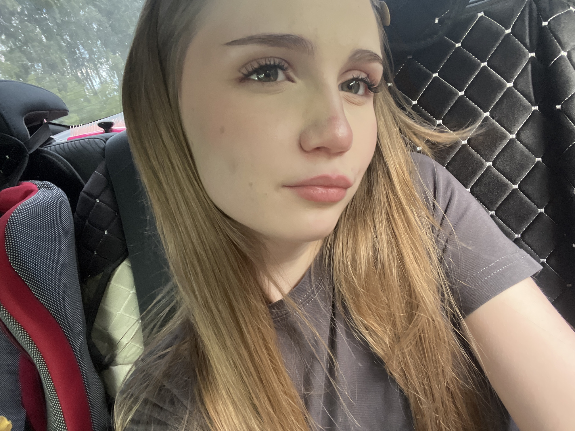 The image shows a close-up, front-facing selfie of a young woman with long blonde hair. She is wearing a gray t-shirt. Her makeup includes eyeshadow and mascara. The background suggests she is in a car; the seat upholstery with a diamond pattern is visible on the right, and a car seat with red and gray accents is on the left. Trees and foliage are visible through the rear window. The image appears to be taken during the daytime, with natural light illuminating the scene. The woman is looking directly at the camera. No specific location in Загородный, Russia is identifiable from the image.