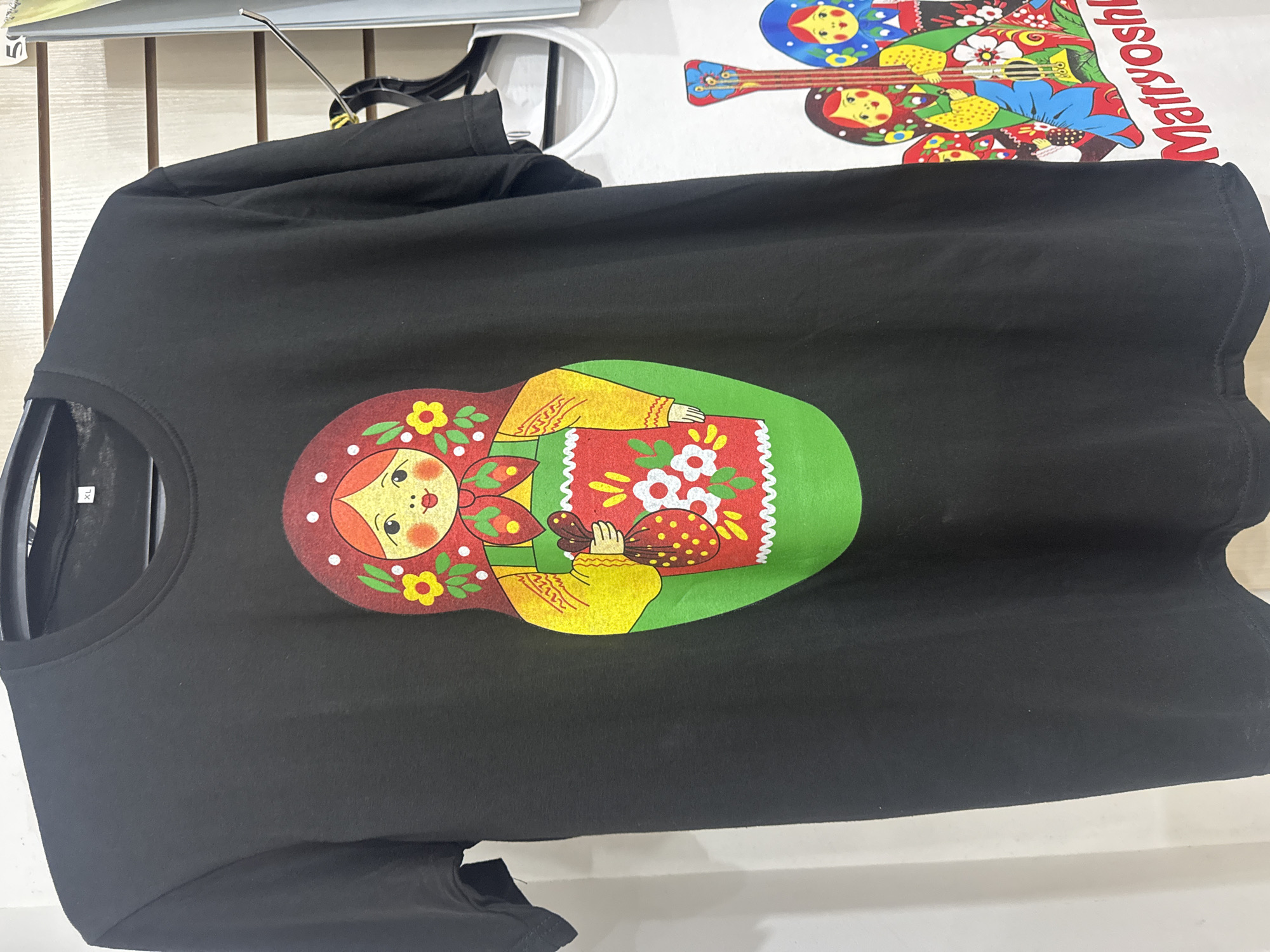 A close-up shot showcases a black t-shirt hanging on a clothes rack. The t-shirt features a vibrant graphic print of a matryoshka doll, a traditional Russian nesting doll, in a cartoonish style. The doll has a red headscarf adorned with white polka dots, rosy cheeks, and a friendly expression. It is dressed in a yellow blouse with intricate red detailing and a red apron decorated with white and red flowers. The doll is holding a small red bag filled with white flowers. The background consists of a slatted wall, commonly found in retail environments. To the right of the black t-shirt, a white t-shirt is partially visible, also displaying a graphic of matryoshka dolls and a guitar, with the word "Matryoshka" written in red lettering. The scene appears to be within a shop or market stall. The lighting is neutral, and the focus is sharp on the t-shirts. There are no individuals visible in the frame. The overall impression is that of merchandise displayed for sale.