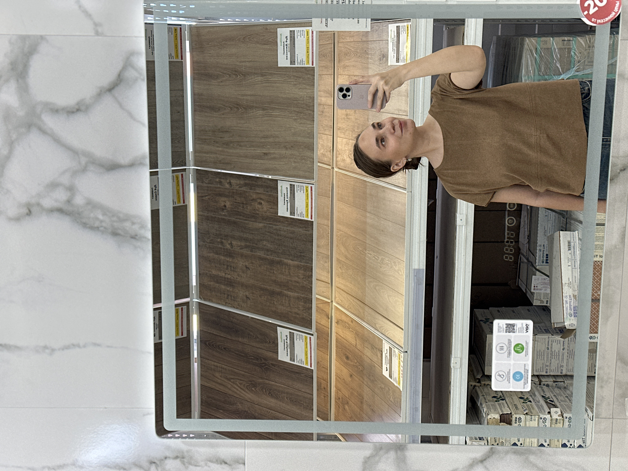 The image shows a woman taking a selfie in a mirror at a store in Chaikovsky, Russia. The mirror reflects a display of different types of wood flooring. In the background, there are boxes of tile flooring, and a small tag indicates "-20% off." The floor in front of the mirror is white marble.