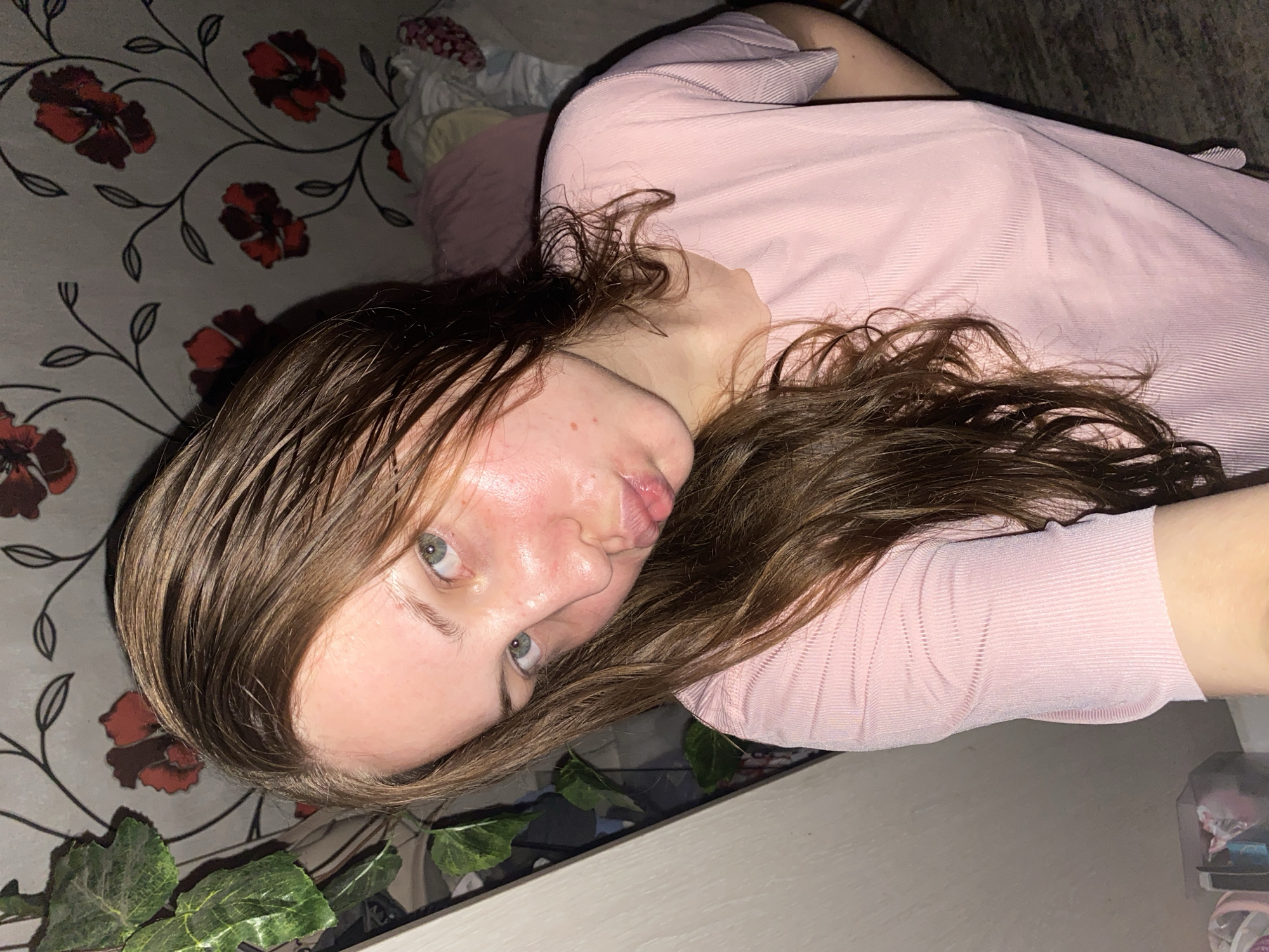 The image shows a young woman with long, brown, wet hair, looking at the camera with her lips puckered. She has light-colored eyes and her face is flushed, with some redness on her cheeks and chin. She is wearing a light pink, ribbed t-shirt. The background consists of floral wallpaper with red and dark red flowers on a grey background. To the left of the woman, there are artificial green leaves. To the right, there are pillows and bedding. The lighting appears to be artificial and warm. The image was taken indoors, likely in a residential setting in the city of Vasyutina, Russia, based on the information provided.