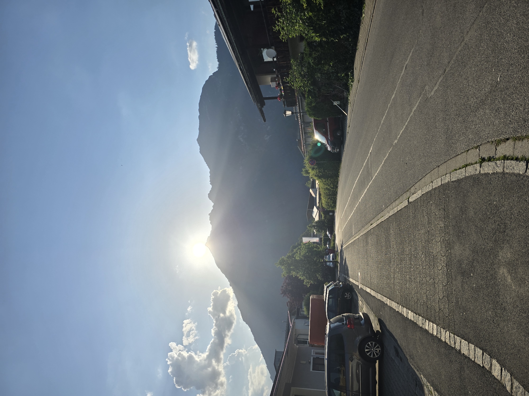The image depicts a street scene in Garmisch-Partenkirchen, Germany, with a mountain range dominating the background. The sun is setting behind the mountain, casting a warm glow and long shadows across the street and surrounding buildings. On the left side of the street, parked cars, including a dark gray minivan and a smaller black SUV, are visible in front of a low-rise building. A sidewalk with a cobblestone border runs alongside the parked cars. The street itself is paved with asphalt and has lane markings. On the right side of the street, a house with a balcony, covered in lush green foliage, is prominent. A red van is parked further down the street, near the house. Trees and more vegetation line the street, contributing to a suburban or semi-rural atmosphere. The sky is a clear blue with a few scattered clouds, some of which are illuminated by the setting sun. The overall impression is one of a peaceful, late afternoon in a picturesque German town.