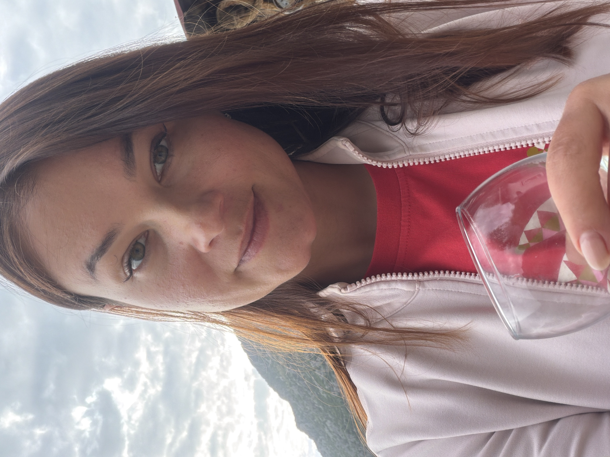 A woman with light brown hair and green eyes is holding a clear glass with a pink and white geometric pattern in front of her. She is wearing a red shirt and a light-colored jacket. The background shows a cloudy sky, mountains, and possibly a body of water. Given the prompt, the location is Donji Orahovac, Montenegro.