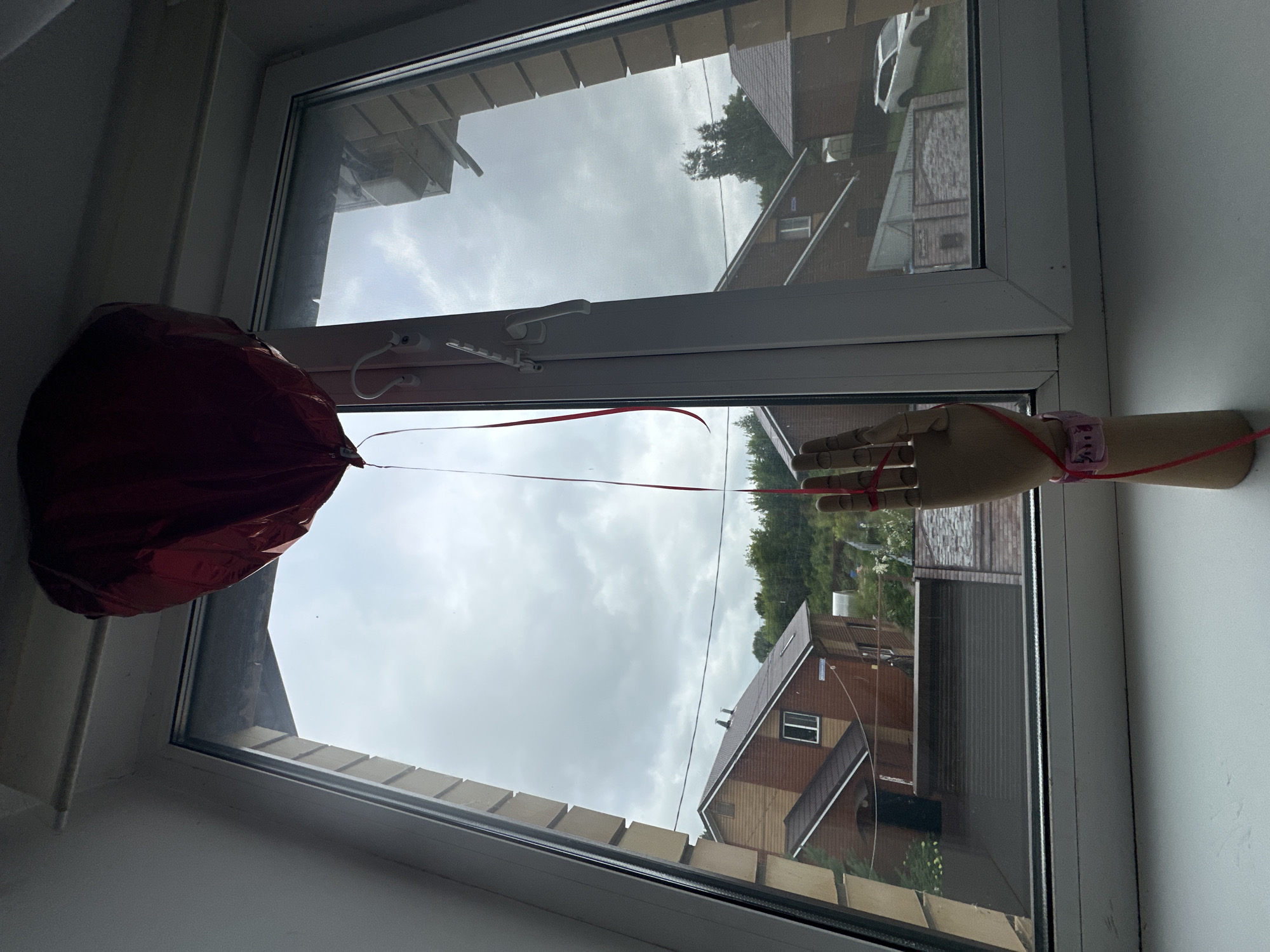 A red heart-shaped balloon is suspended by a thin ribbon, which is tied to the fingers of a wooden mannequin hand. The mannequin hand is positioned on a white windowsill. The ribbon also wraps around the fingers of the mannequin hand and extends down to a pink watch on its wrist. The scene is viewed through a window, looking out onto a cloudy sky and a residential area with brick houses and trees. A white car is visible parked in front of one of the houses.