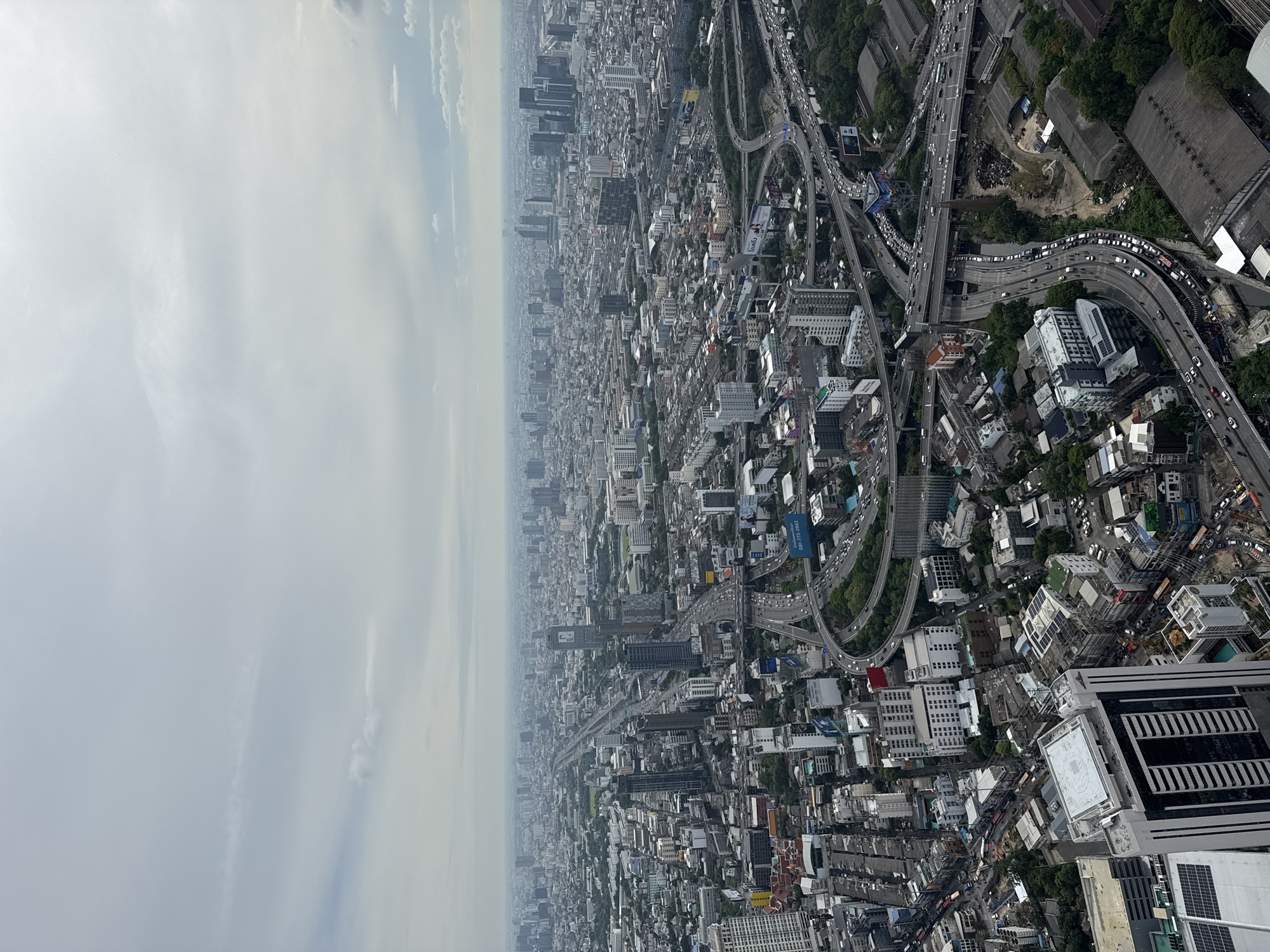 The image shows an aerial view of Bangkok, Thailand. The scene is dominated by a dense urban landscape with numerous buildings of varying heights and architectural styles. A complex network of elevated highways and roads is visible, congested with vehicular traffic. The sky is overcast and hazy. Vegetation is scattered throughout the urban environment, primarily around residential areas. The photo shows a sprawling cityscape with a mix of residential, commercial, and industrial zones.