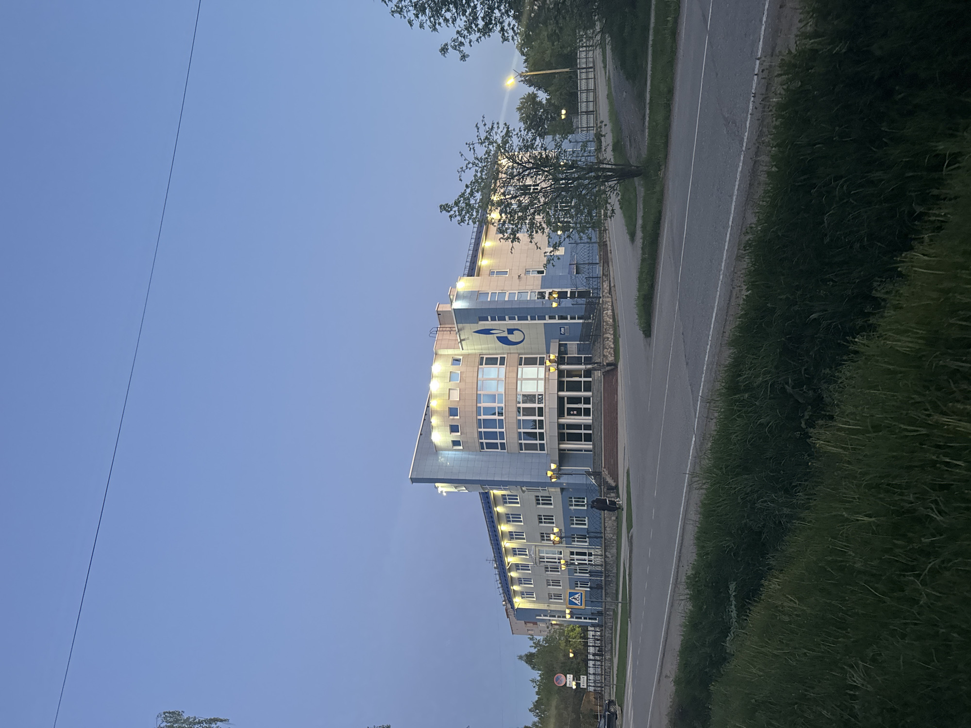 The image presents a view of the Gazprom building in Berezovsky, Russia, during the evening hours. The building is a multi-story structure, featuring a blue and white facade with numerous windows. The Gazprom logo is prominently displayed on the building's exterior. The building is adjacent to a street and sidewalk. Vegetation, including trees and shrubs, is visible along the roadside. The sky is a clear blue gradient, transitioning into darker shades. Street lighting illuminates the surrounding area. No individuals are clearly visible in the image.