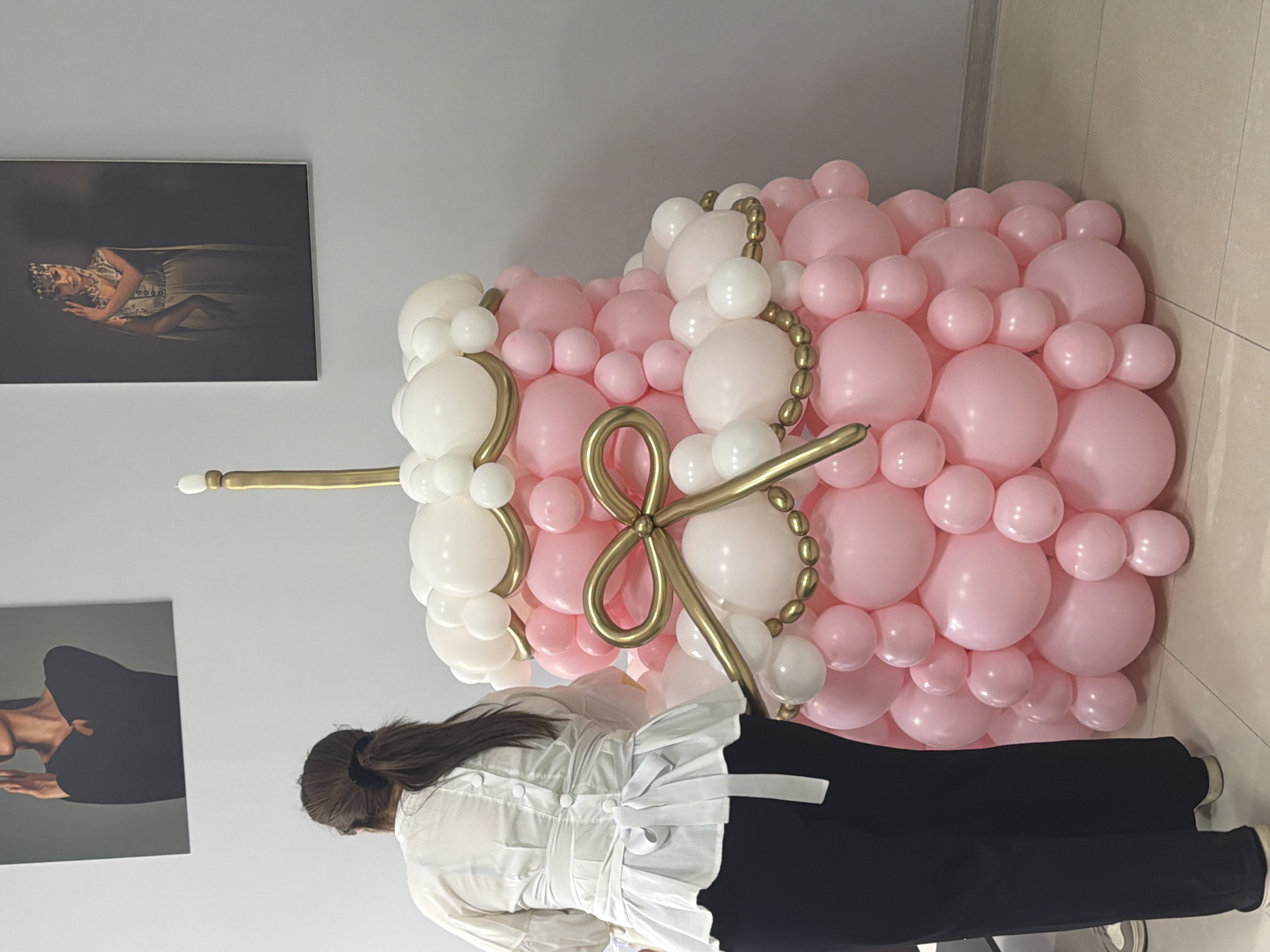 A scene captured indoors in Yerevan, Armenia, features a woman with dark hair pulled back, wearing a white blouse and black pants, positioned in front of a large balloon sculpture resembling a cupcake. The sculpture is primarily pink and white, adorned with golden balloon accents. Two framed photographs hang on the wall behind the woman and the sculpture; one shows a person lying down, while the other depicts a person in a dark outfit. The floor is tiled, and the lighting appears to be artificial. The woman seems to be interacting with or near the balloon sculpture.