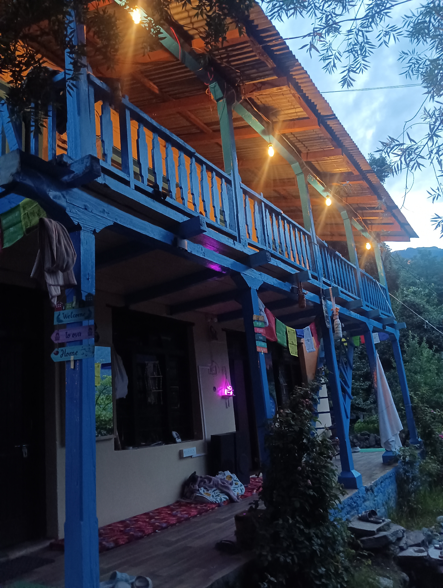 The image depicts a rustic, multi-story wooden structure, likely a guesthouse or homestay, in a hilly, green environment. The building features a prominent blue-painted balcony with a railing, supported by blue wooden pillars. String lights with warm yellow bulbs are visible along the underside of the balcony roof, and a few small prayer flags in various colors are hung from the railing and pillars.
A sign with directional arrows is attached to one of the front pillars, reading "Welcome," "to our," and "Home" in a whimsical font. Clothing, possibly towels or garments, are draped over the balcony railing and pillars. The lower level of the building has a dark-colored door and a window with a view of greenery. The ground in front of the building is a wooden deck, with sleeping mats and cushions laid out, suggesting an outdoor living or resting area. There are also some sandals on the ground.
The background shows lush green trees and vegetation, with a mountainous landscape visible in the distance, consistent with the topography of Old Manali, India. The lighting suggests it is either dusk or early evening, with the sky appearing overcast. No people are clearly visible in the image, but the setup implies a welcoming and lived-in space. The overall aesthetic is one of a relaxed, natural, and possibly bohemian retreat.