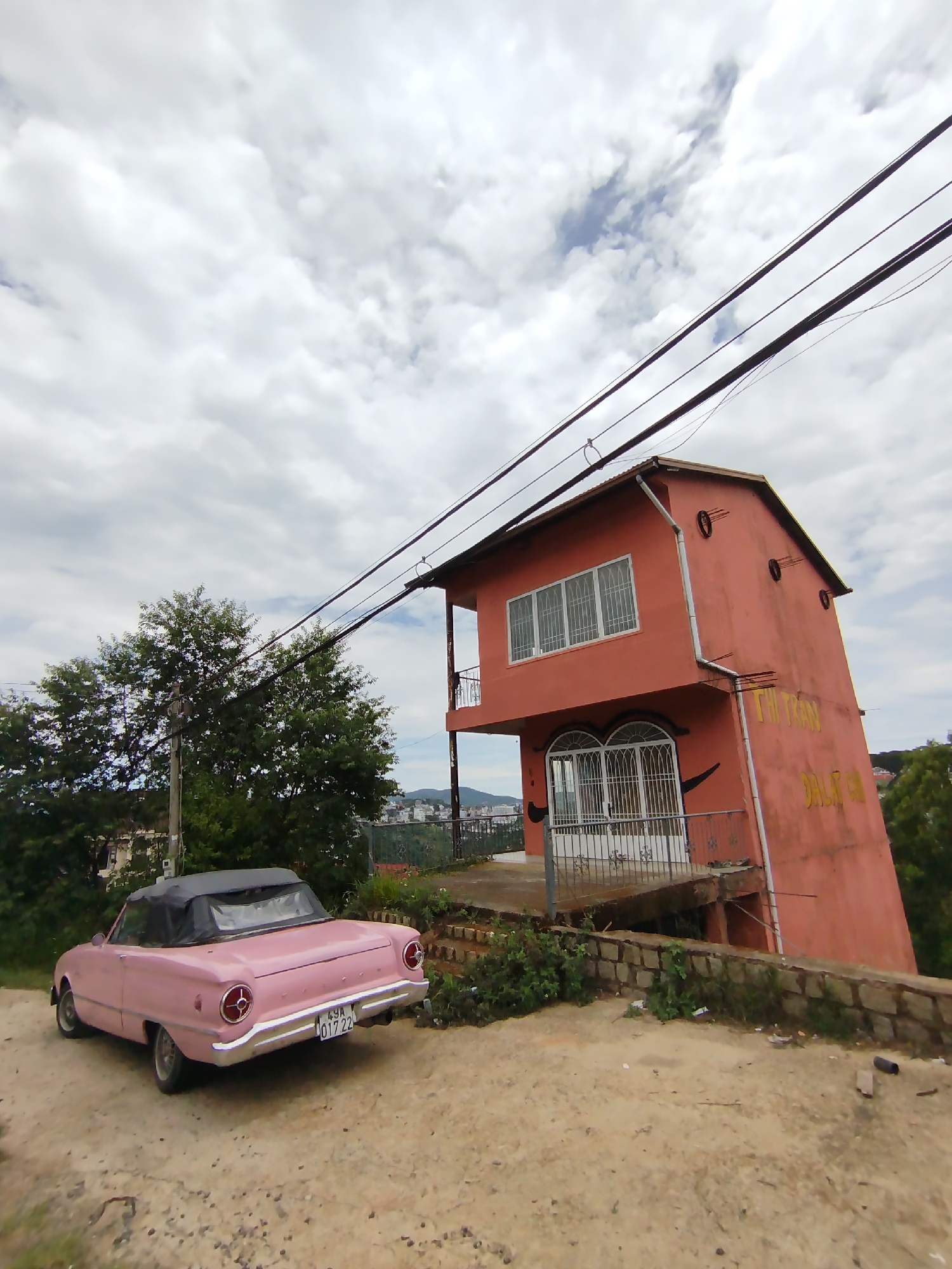 The image shows a pink vintage convertible car parked on a dirt road in front of a leaning, two-story, pink building. The building features a balcony, arched windows on the ground floor, and rectangular windows on the upper floor. Yellow writing is visible on the side of the building. Power lines run across the top of the frame, connecting to the building. Behind the building, a cityscape is visible in the distance. A tree stands to the left of the car. The sky is overcast with white clouds. The car has a black convertible top. The license plate is visible but not easily legible. The scene appears to be in Da Lat, Vietnam, based on the architectural style of the building.