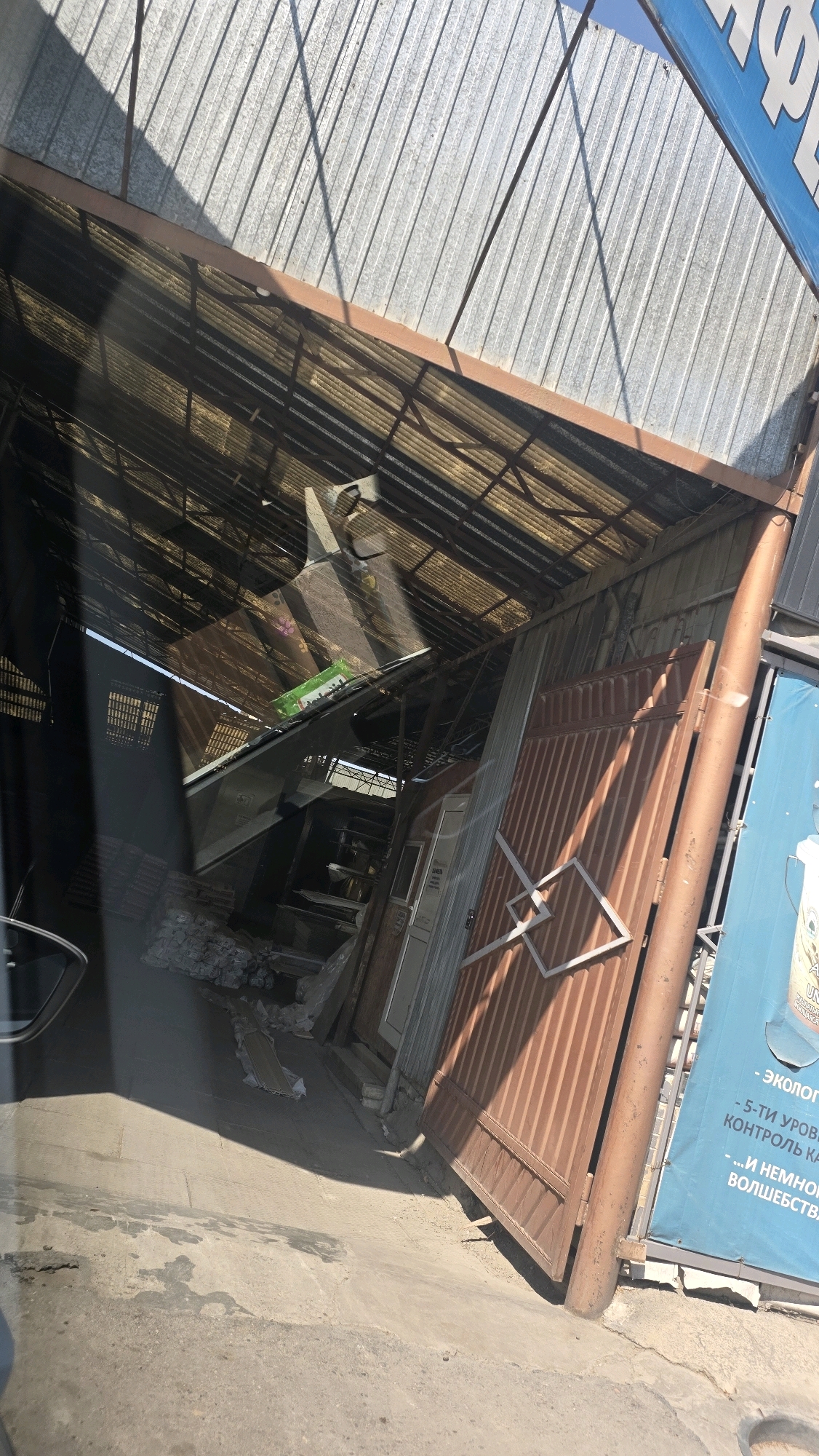 The image shows an outdoor scene with a corrugated metal roof supported by beams, appearing somewhat dilapidated. A large, brown metal gate is partially open, revealing an interior space with various items. Among these are what appear to be building materials, possibly stacks of insulation or plasterboard, and other assorted objects. A green package is visible on a sloped surface within the structure. The ground is concrete and appears uneven, with some water stains. A blue banner with text in Cyrillic is attached to a wall on the right side of the image. The overall impression is of a storage area or an informal commercial space, possibly in a developing part of Tokmok City, Kyrgyzstan. The lighting suggests it is daytime, with shadows cast by the overhead structure.