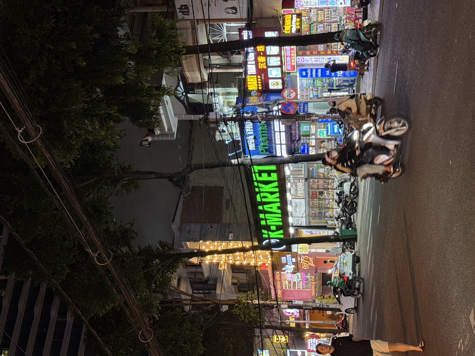 The image depicts a street scene at night in Nha Trang, Vietnam. A street is paved with asphalt, with visible lane markings. On the right side of the street, a sidewalk is lined with shops and businesses featuring bright neon signs. One prominent sign reads "K-MARKET" in large green lettering. Other businesses appear to include a pharmacy, indicated by a sign that says "Pharma" and a red sign with Chinese characters. There are trees lining the street, and power lines are strung overhead. Several motorcycles are present on the street, with two in the foreground appearing to be in motion. One motorcycle is ridden by two people, and another is ridden by a single person. A white car is parked on the left side of the street. A pedestrian, a man wearing a dark shirt and light shorts, is walking on the left side of the street towards the camera. The overall environment is that of a busy commercial street at night.