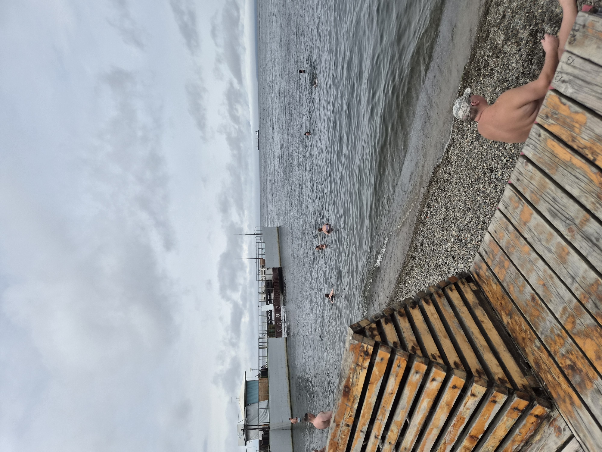 The image depicts a body of water, likely a bay or sea, with a pier structure in the middle ground. Several people are visible swimming and wading in the water. The foreground features a wooden deck and a pyramid-shaped wooden structure. In the background, a cargo ship is visible on the horizon, and a cloudy sky dominates the upper portion of the image. The scene suggests a recreational area, possibly a public beach or a swimming spot, with a weathered appearance to the wooden elements. The location is identified as Vladivostok, Russia, a port city known for its coastal landscape and maritime activity.