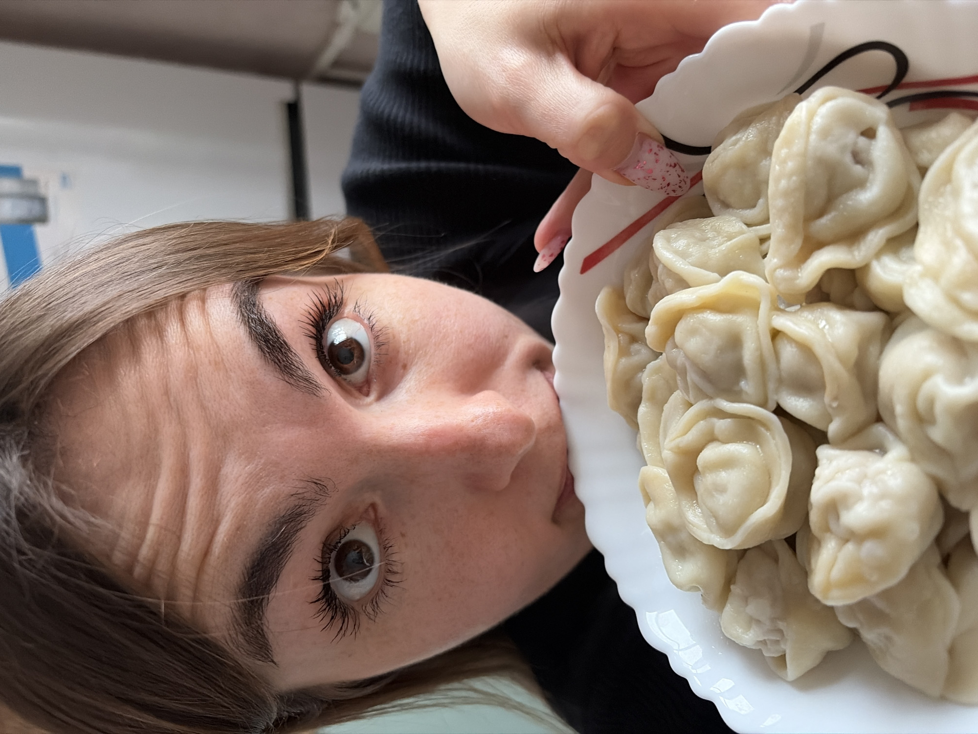 A woman is holding a white plate full of pelmeni, a type of dumpling. Her left eye is visible, and she appears to be looking at the viewer. Her left hand is holding the plate, and the nails are painted. The background is blurry. The location is not discernible from the image. The photo does not contain evidence of Izhevsk.