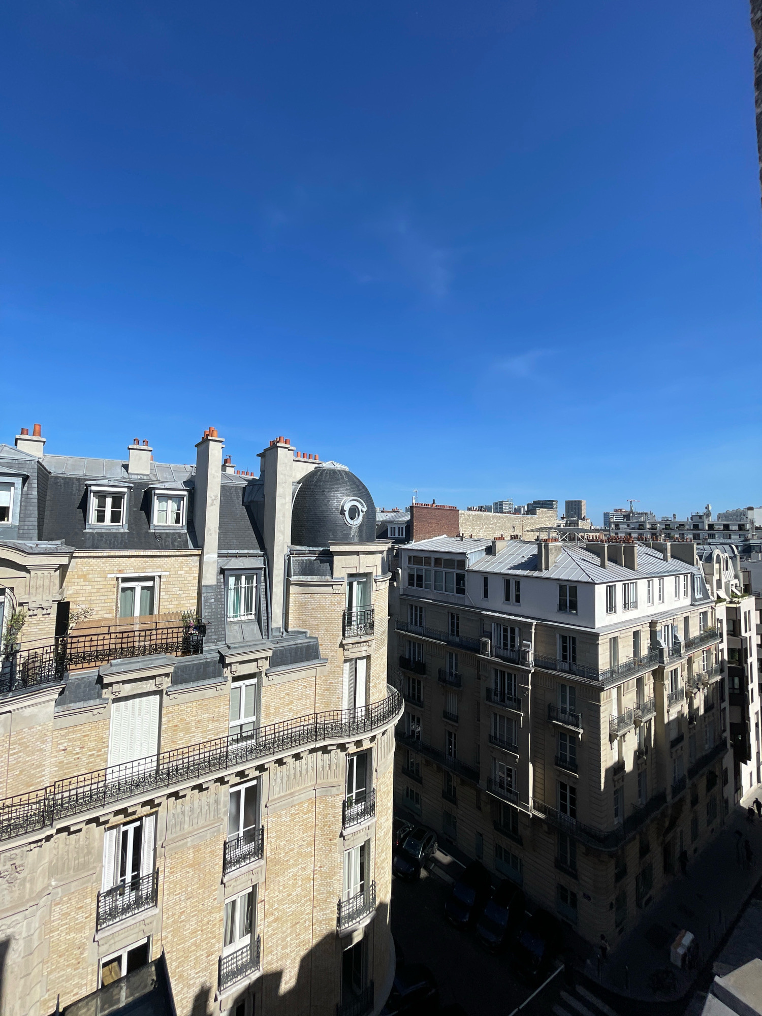 This is an aerial shot of Parisian buildings under a clear blue sky. The foreground features a close-up of ornate apartment buildings with light-colored brick facades, mansard roofs covered in dark grey slate, and numerous dormer windows. Black wrought-iron balconies with intricate designs line the upper floors. A distinct domed structure with a circular window is visible on the roof of one building.
Beyond these buildings, a cityscape unfolds with a mix of architectural styles, including modern high-rises and older structures. Some buildings have grey, peaked roofs, while others exhibit flat roofs. The street level is visible in the lower right quadrant, showing a road with parked cars and a few pedestrians. Sunlight casts strong shadows on the buildings, indicating a bright, sunny day. The overall impression is of a dense urban environment in Paris, France.