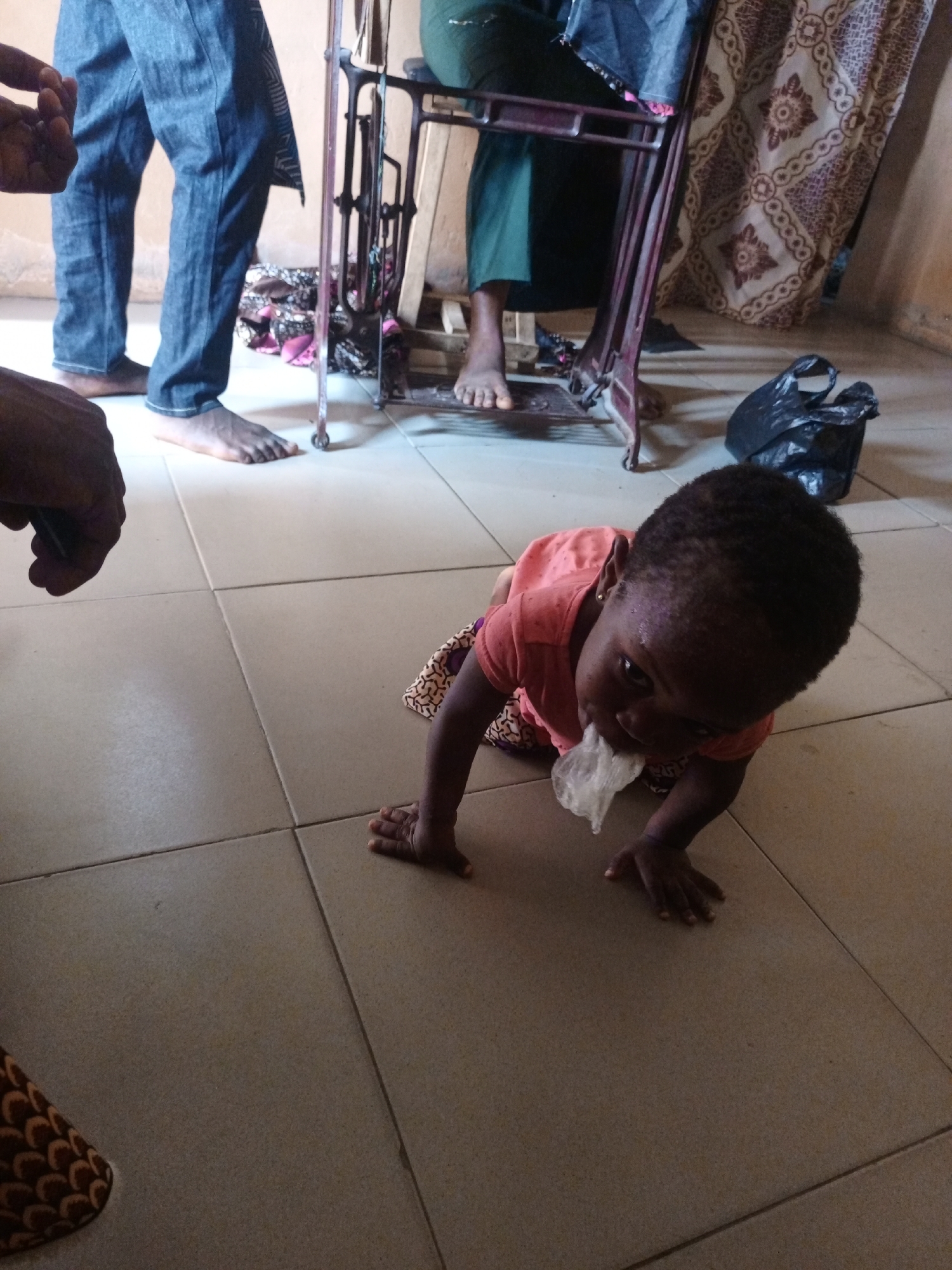 A child crawls on the tiled floor of a room. The child is dark-skinned, wears an orange shirt, and has a small piece of clear plastic in its mouth. Several adults are standing in the background, near a sewing machine. All individuals are barefoot. A black plastic bag sits on the floor in the background. The location appears to be indoors, potentially a home, in Unknown, Unknown.