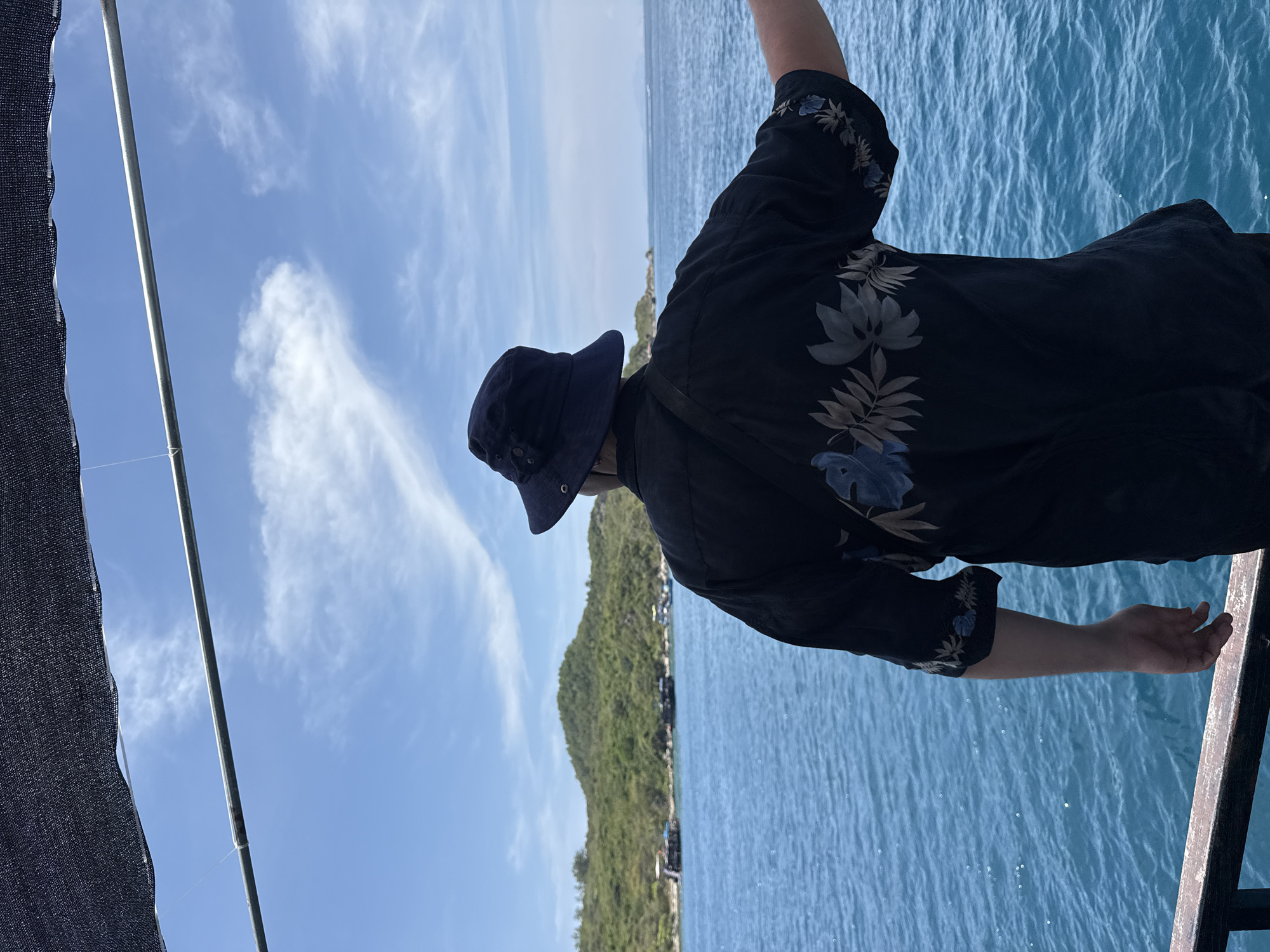 A person stands on a boat in Nha Trang, Vietnam, next to a railing and overlooking the ocean. The person wears a dark hat and a patterned dark shirt. In the background, land covered with green foliage is visible under a partly cloudy blue sky.