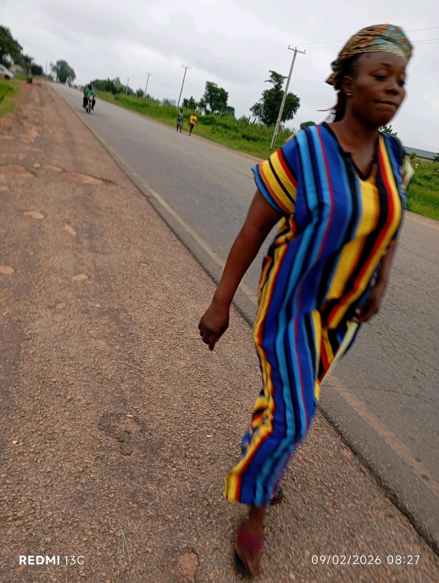 A full-body, slightly blurry, medium shot captures a woman walking along the shoulder of a paved road. She is wearing a colorful, vertically striped dress in shades of blue, yellow, red, and black, and a patterned headscarf. The woman is in motion, her body angled to the right of the frame, and her face is turned away from the camera, giving her a slightly blurred appearance. The road is in the foreground, with a rough, gravelly shoulder on the left and a paved surface with a yellow line on the right. In the background, the road continues into the distance, with green vegetation on either side. Two figures, possibly children, are walking further down the road on the right side, and a motorcycle with a rider is approaching from the left. The sky is overcast, suggesting a cloudy day. The timestamp "REDMI 13C 09/02/2026 08:27" is visible in the bottom left and right corners of the image, indicating the device and date/time of capture. The scene appears to be in Shendam, Nigeria, a town known for its rural setting and agricultural activities.
