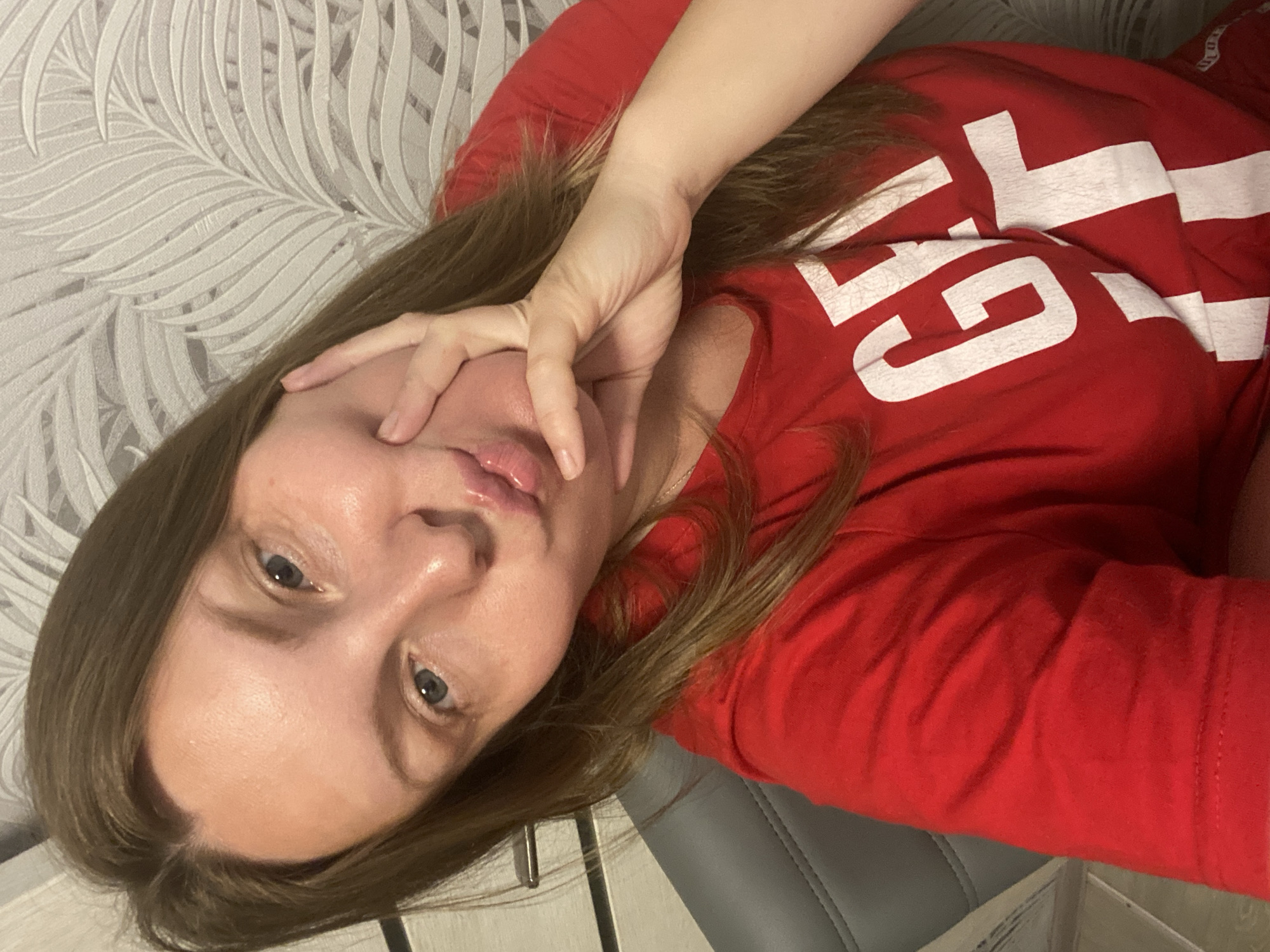 A selfie-style, medium shot shows a woman with shoulder-length brown hair and blue eyes, making a kissing face with her right hand touching her cheek. She is wearing a red t-shirt with white lettering that reads "GET IT." The background consists of a light gray patterned wallpaper and a portion of a gray chair. The image appears to be taken indoors, likely in a residential setting, but there is no specific information to indicate a location in the city of Noviy, Russia.