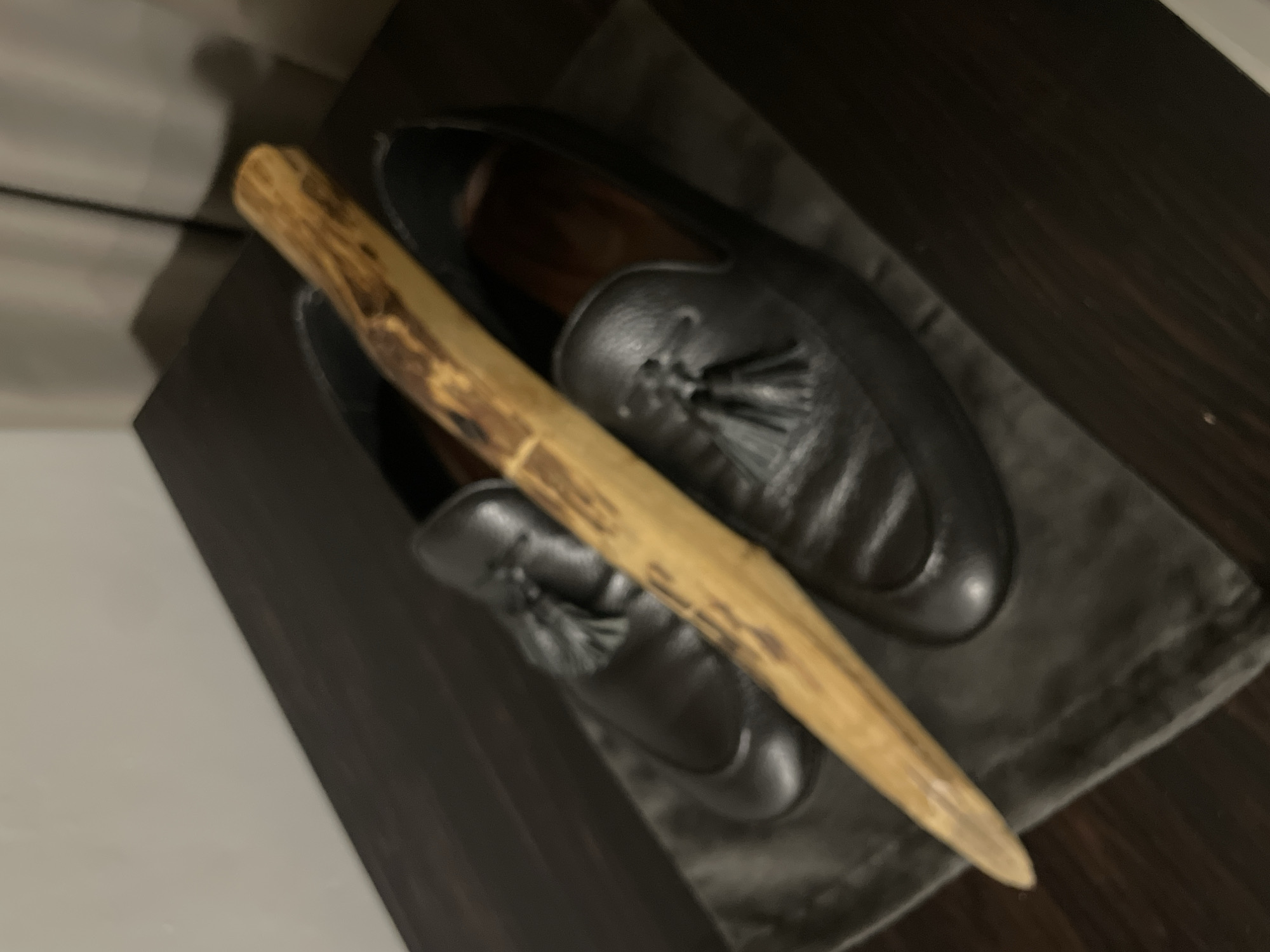 This media file is a close-up, blurry shot of a pair of black leather loafers with tassels. A slender piece of light-colored wood, possibly a twig or a dowel, lies across the top of the shoes. The shoes are resting on a dark gray cloth, which in turn is placed on a dark wooden surface. The background is out of focus, showing hints of a light-colored wall and a shadow. The lighting suggests an indoor setting, possibly dim. The overall impression is of still life, with the objects arranged deliberately. There is no indication of people, an activity or event, time of day, weather, or emotions. No text is visible. The location context of Sakhu, Thailand is not visually evident in the image.