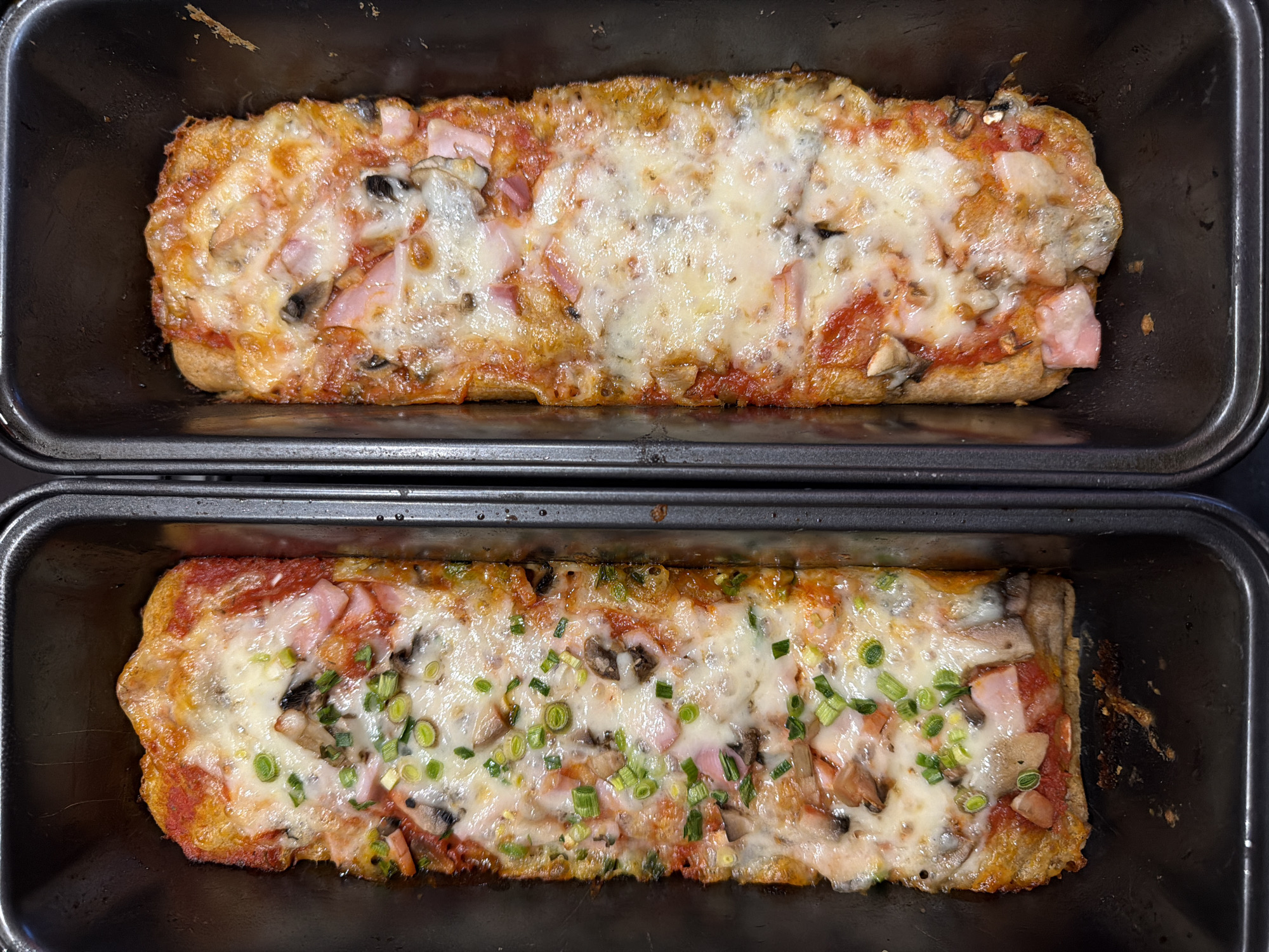 Two rectangular black baking pans sit side-by-side. Each pan contains a baked rectangular pizza or bread topped with tomato sauce, cheese, ham, mushrooms, and possibly other toppings. The bottom pizza has sliced green onions sprinkled on top. The location in Varna, Bulgaria, is not determinable from the visual information.