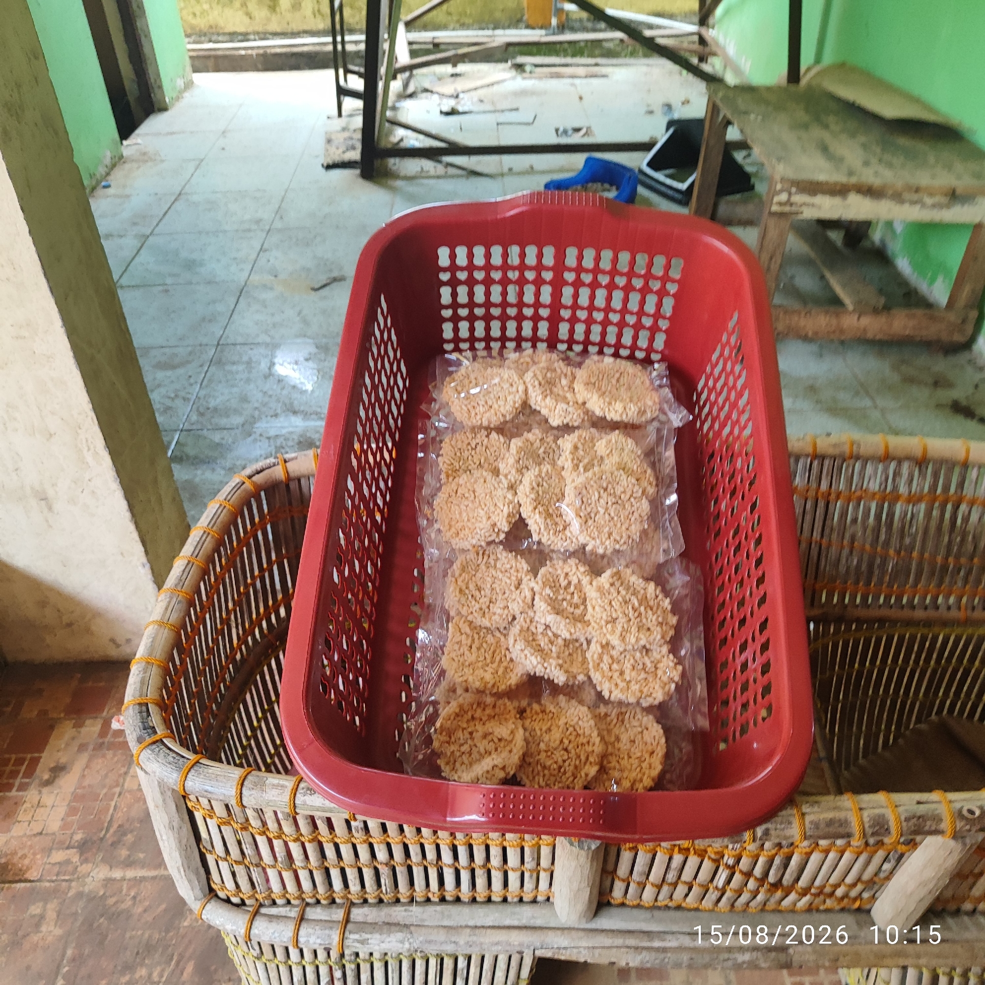 In Pembaharuan, Indonesia, an outdoor scene captures a red plastic basket filled with individually packaged, round, light brown crackers. The basket rests on top of a larger, cylindrical woven basket made of bamboo and tied with orange string. Behind the baskets, a concrete floor is visible, with some scattered debris and a green wall on the left. In the background, a makeshift wooden table and metal shelving unit are present, suggesting a storage or market area. A date and time stamp of "15/08/2026 10:15" is visible in the bottom right corner of the image.