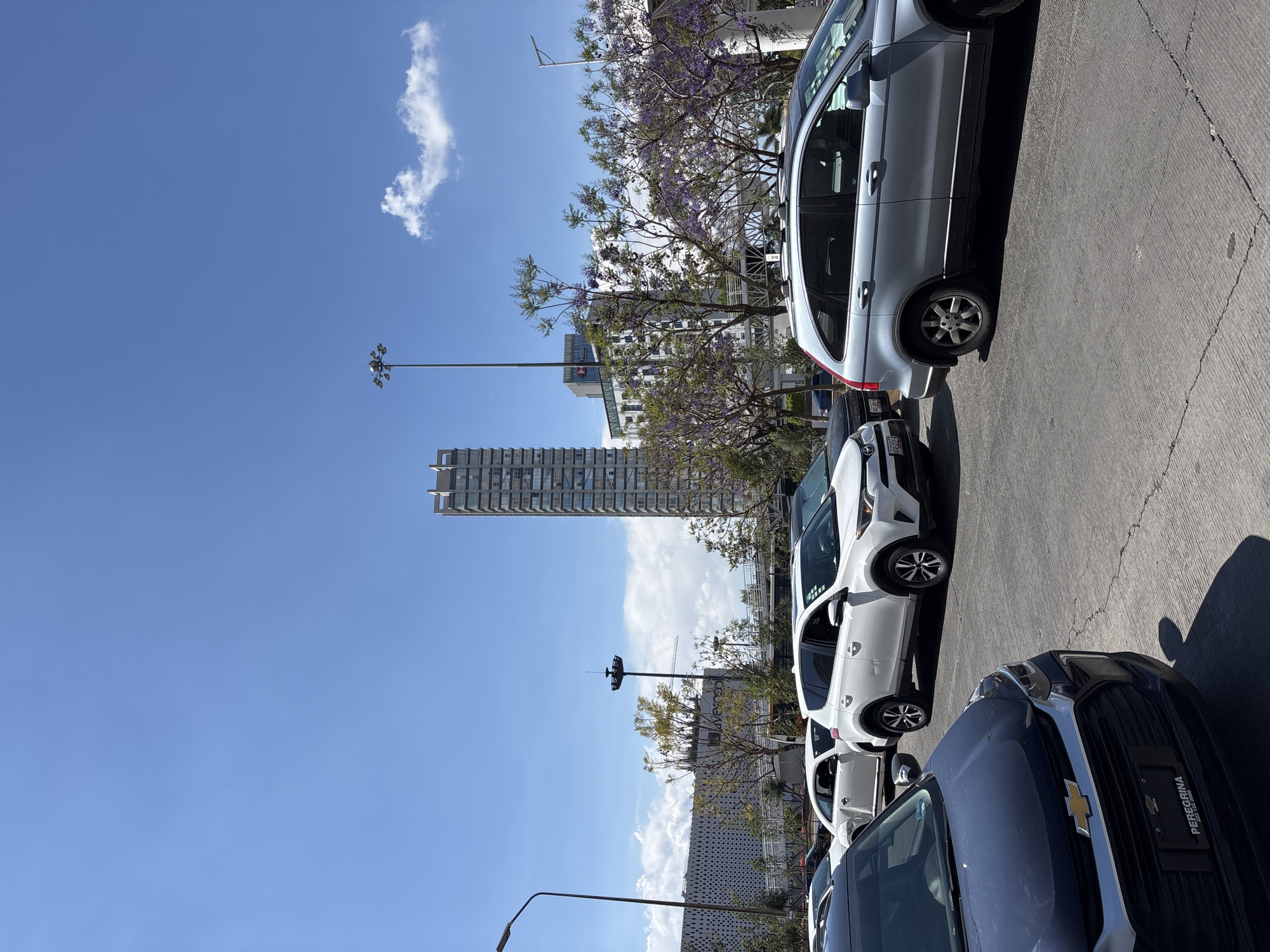 The image depicts a street scene in Puebla, Mexico, characterized by a row of parked cars along the right side. The vehicles, varying in color and model, include a gray SUV, a white SUV, and a dark blue Chevrolet. Buildings and a high-rise structure are visible in the background, along with flowering trees with purple blossoms. A clear, blue sky occupies the majority of the frame, featuring a singular cloud formation. Streetlights line the edge of the street.