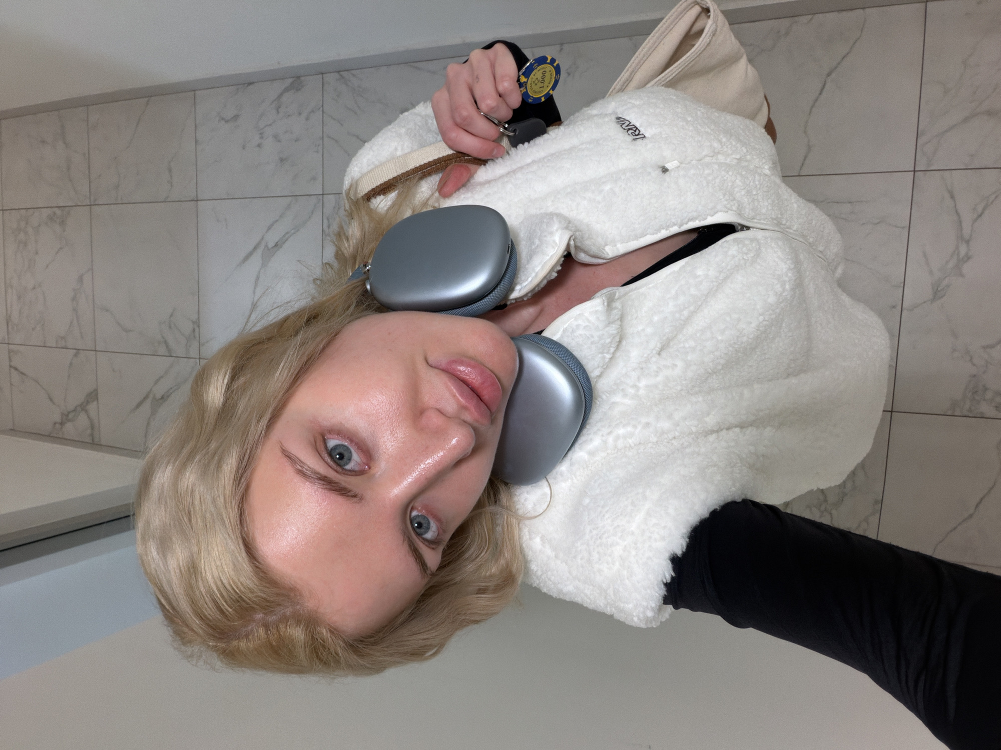 A fair-skinned woman with blonde hair and blue eyes is posing for a selfie. She is wearing a white, sherpa-lined jacket with a hood and a black long-sleeved shirt. Around her neck are silver headphones. She is holding keys and a blue and yellow casino chip in her left hand. She is wearing a white shoulder bag.
The background is a wall with marble-patterned tiles. The location is indeterminate, but it is not possible to confirm it is in Belgrade, Serbia based solely on the visual information provided.
