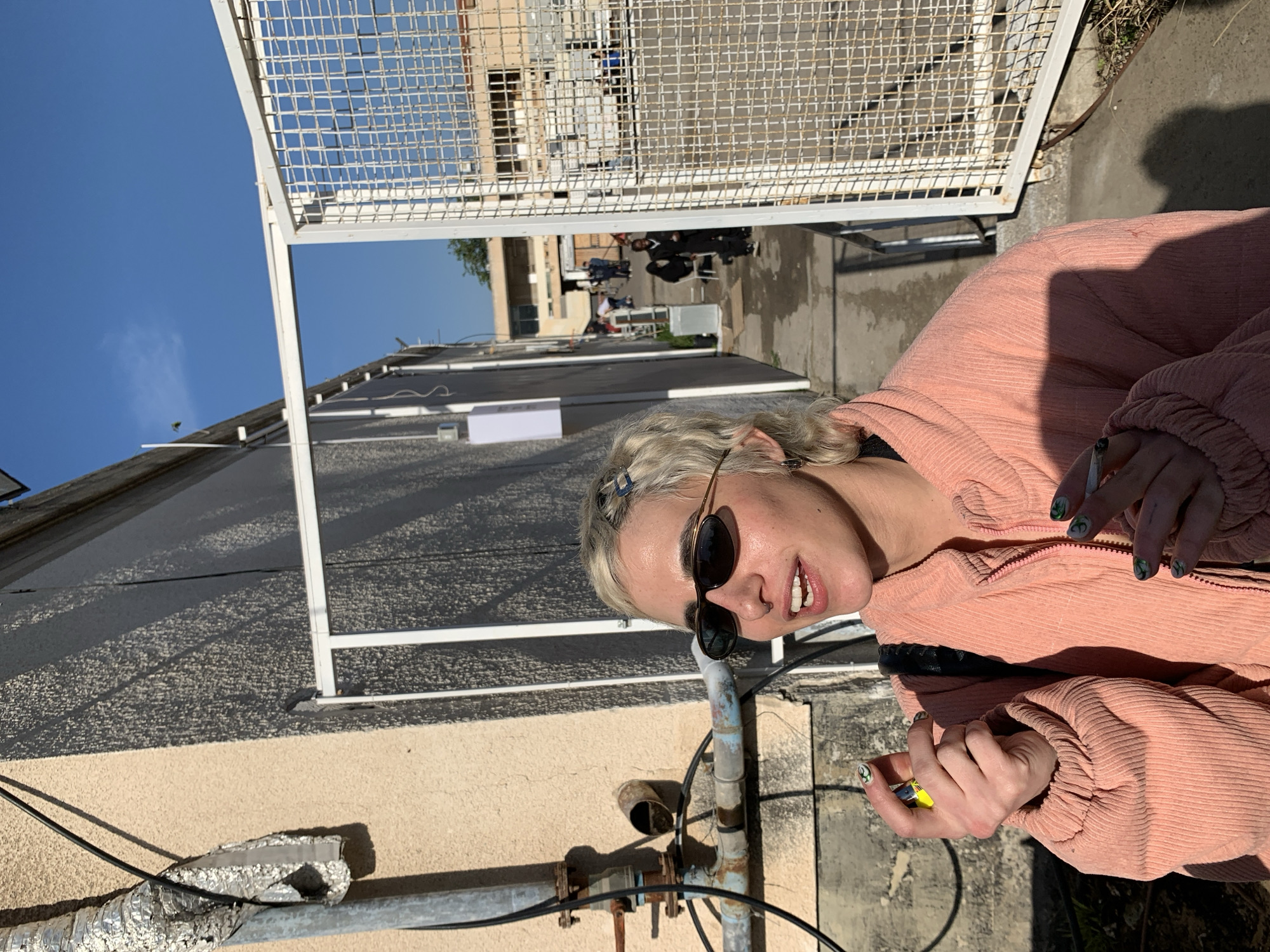 The image depicts a young adult, with short bleached blonde hair and dark oval sunglasses, standing outdoors in Tel-Aviv, Israel. They are wearing a salmon-pink corduroy zippered jacket. Their left hand holds a hand-rolled cigarette, while their right hand holds a yellow disposable lighter, poised as if to light the cigarette. Their nails are painted dark green with white stylized designs. The individual's mouth is slightly open, and their head is turned to the left.
To the left, a light-colored, textured stucco wall features a corroded metal pipe system with exposed wiring and a silver foil-wrapped ventilation duct. Bright sunlight casts sharp shadows on this wall. To the right, an open white metal gate with a grid pattern leads into a narrow outdoor passageway or alleyway.
Beyond the gate, a pathway stretches into the distance, where several individuals are visible standing or seated, indicating a public or communal space. In the background, utilitarian or older buildings with various roof types are present under a clear blue sky. The ground in the pathway appears to be concrete or asphalt, with some unevenness and debris.