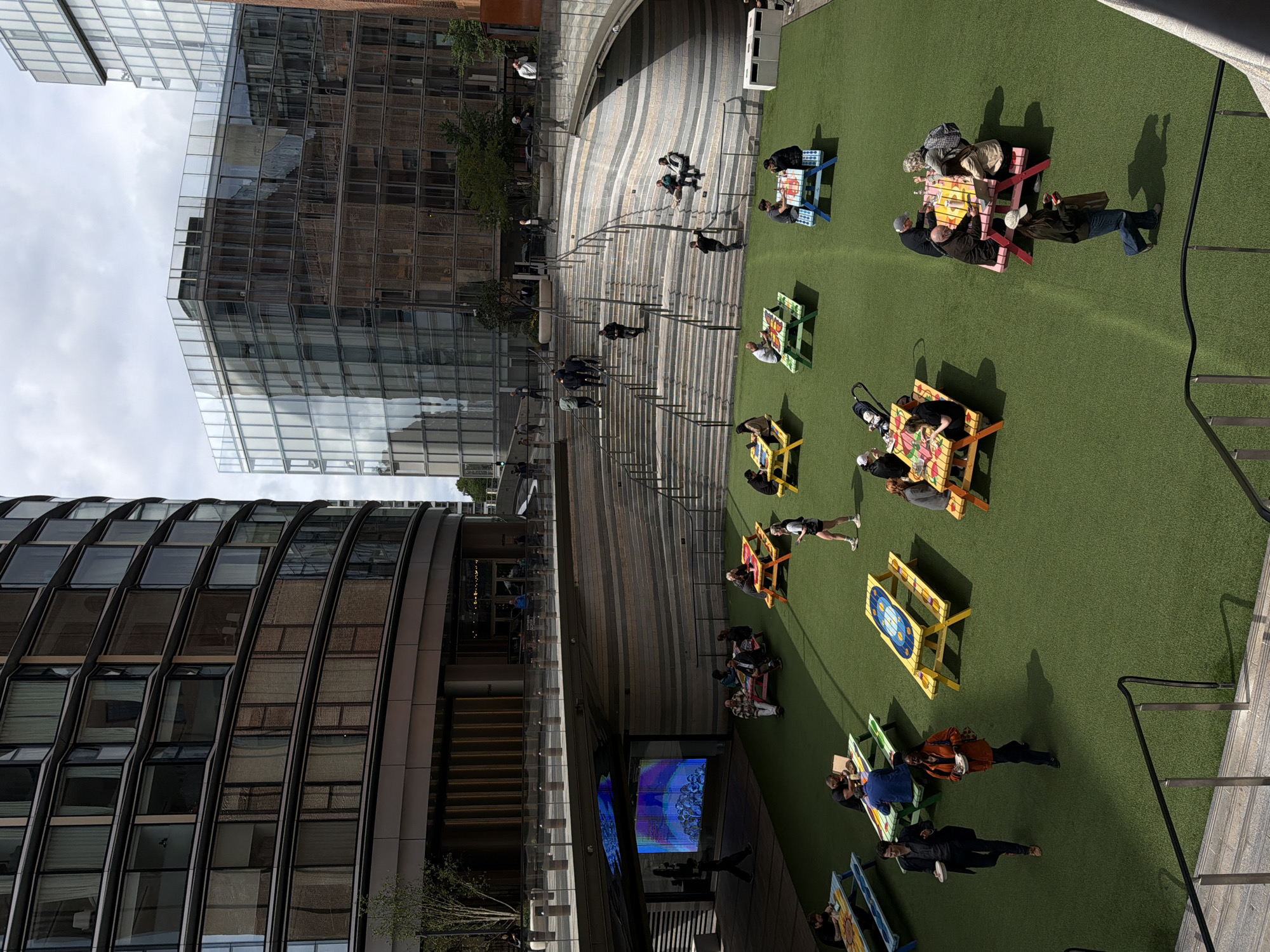 The image shows an elevated view of an outdoor urban space, likely in the City of London, United Kingdom, given the modern architecture. A green, artificial turf area is populated with numerous colorful picnic tables, many occupied by people. Individuals are sitting, standing, and walking in the area, some appearing to be eating or engaged in conversation. A large digital screen is visible near the base of one of the buildings. Several sets of stairs connect different levels of the space, and people are using them to move around. The modern buildings feature glass and metal facades. The sky is overcast.