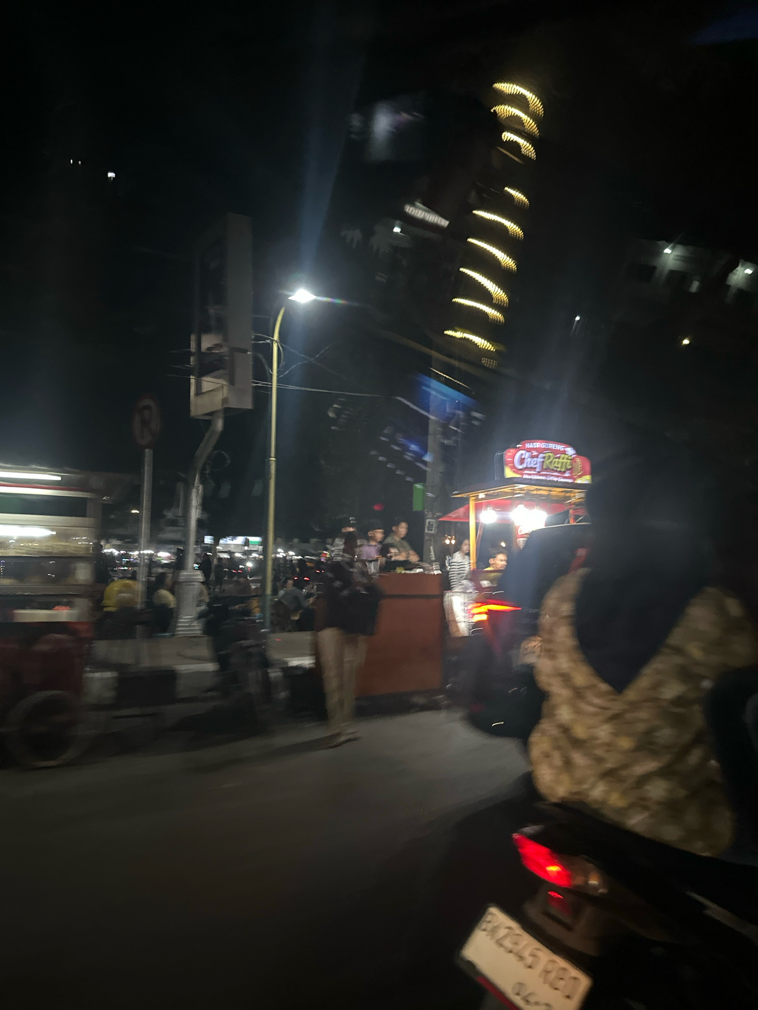 The image captures a nighttime street scene in the City of Binjai, Indonesia. A food stall with a prominent "Chef Raffi" sign is visible, suggesting it is a culinary establishment. The street is illuminated by streetlights and the lights from the food stall, creating a vibrant atmosphere. Several people are present, some appearing to be customers or vendors at the stall, while others are on motorcycles, implying a bustling urban environment. A traffic light and a "no parking" sign are also discernible, indicating the presence of traffic regulations. The overall impression is that of a lively nighttime commercial area in a city.