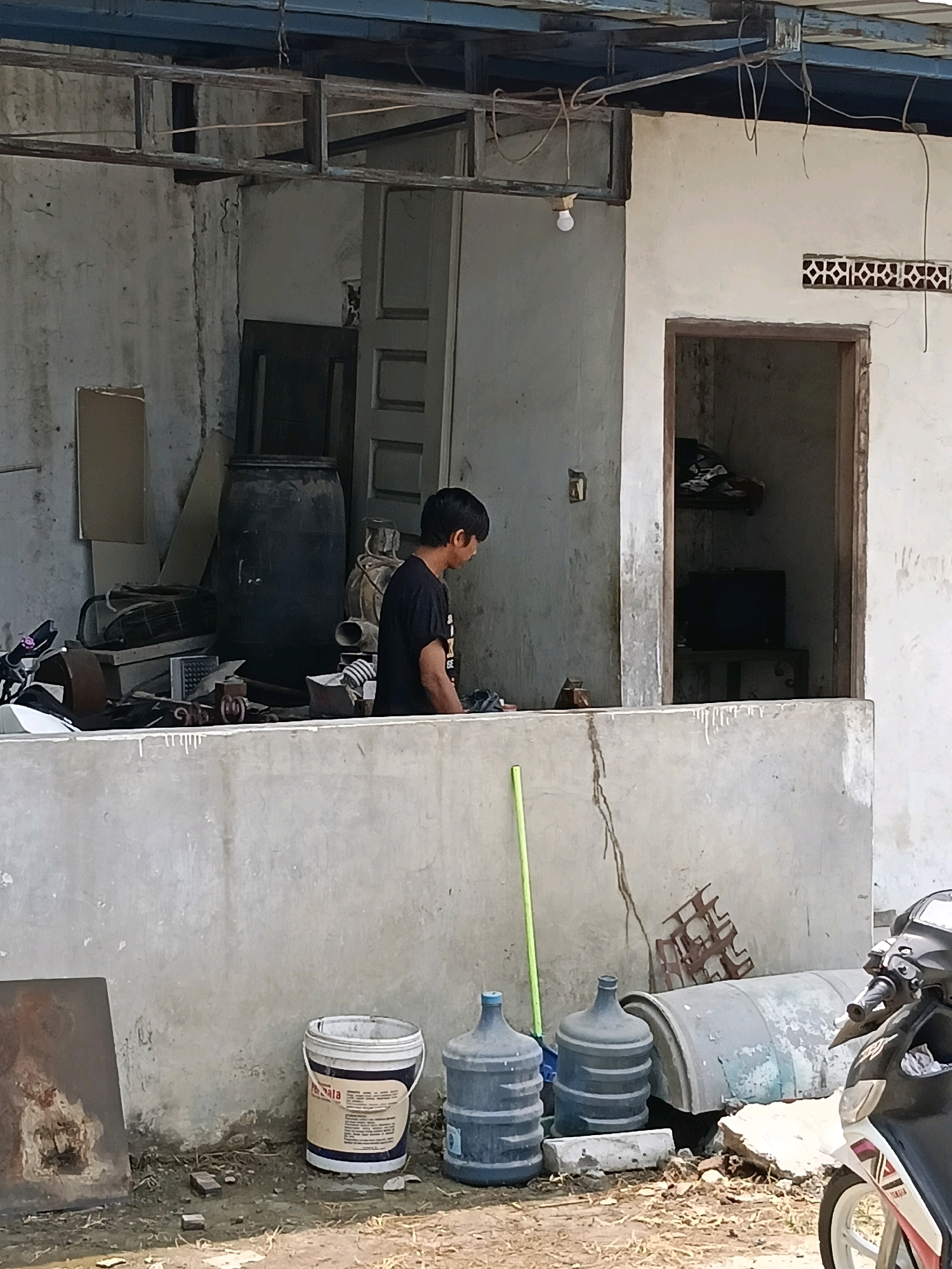 The image depicts an outdoor scene in what appears to be a workshop or storage area in Belimbing, Indonesia. A young man wearing a black t-shirt is standing near a large black barrel, looking downwards as if engaged in some task. The environment is somewhat cluttered, with various objects visible including what looks like scrap metal, old machinery parts, a motorcycle in the foreground to the right, and several large plastic water containers. A section of a concrete wall runs across the middle of the image, separating the foreground from the background where the man and the barrel are situated. The building in the background has a weathered, unfinished look with exposed beams and rough walls. The ground is unpaved and appears to be dirt and debris. The lighting suggests it is daytime, with sunlight casting shadows.
