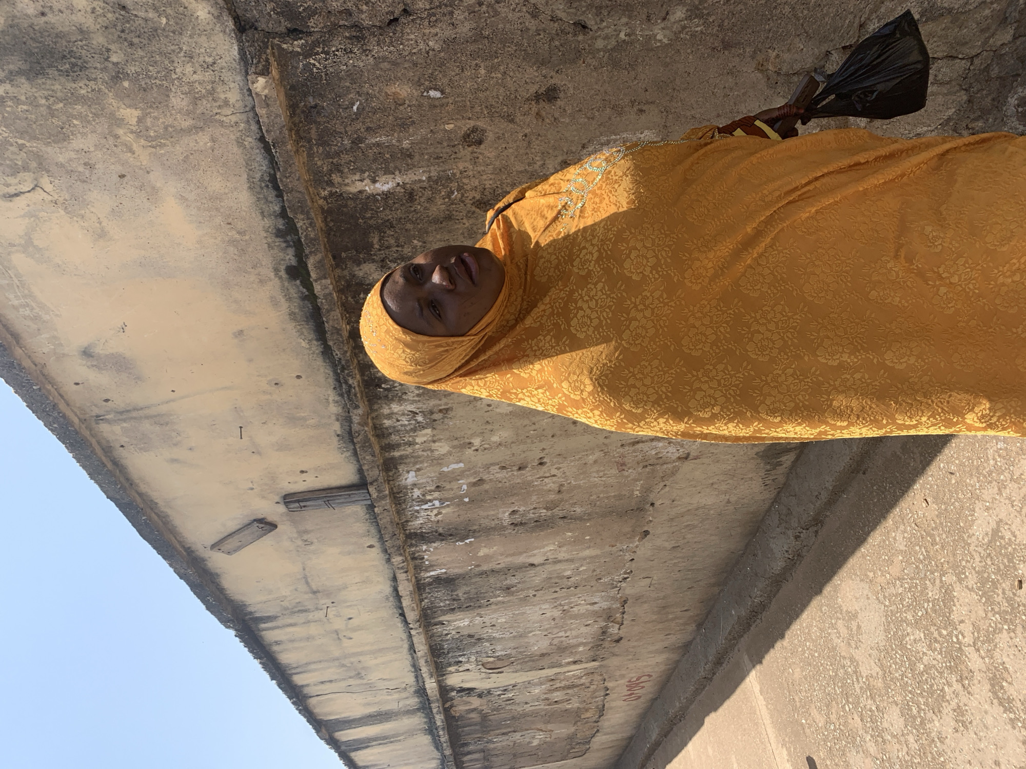 A woman wearing an orange hijab and dress stands beside a textured, weathered wall under a clear blue sky. The wall, which appears to be made of concrete or stucco, is a light yellow color and shows signs of age, including cracks and peeling paint. The woman is looking to her right and holding a black plastic bag in her right hand. A portion of the ground, which is a dusty, light brown color, is visible in the foreground, with a shadow cast across it. The image appears to have been taken outdoors during the daytime. The wall has some markings on it, including what looks like "07045" in red paint. There are also some wooden planks and nails embedded in the upper part of the wall. This location is likely in Jos, Nigeria, given the style of the wall and the appearance of the ground.