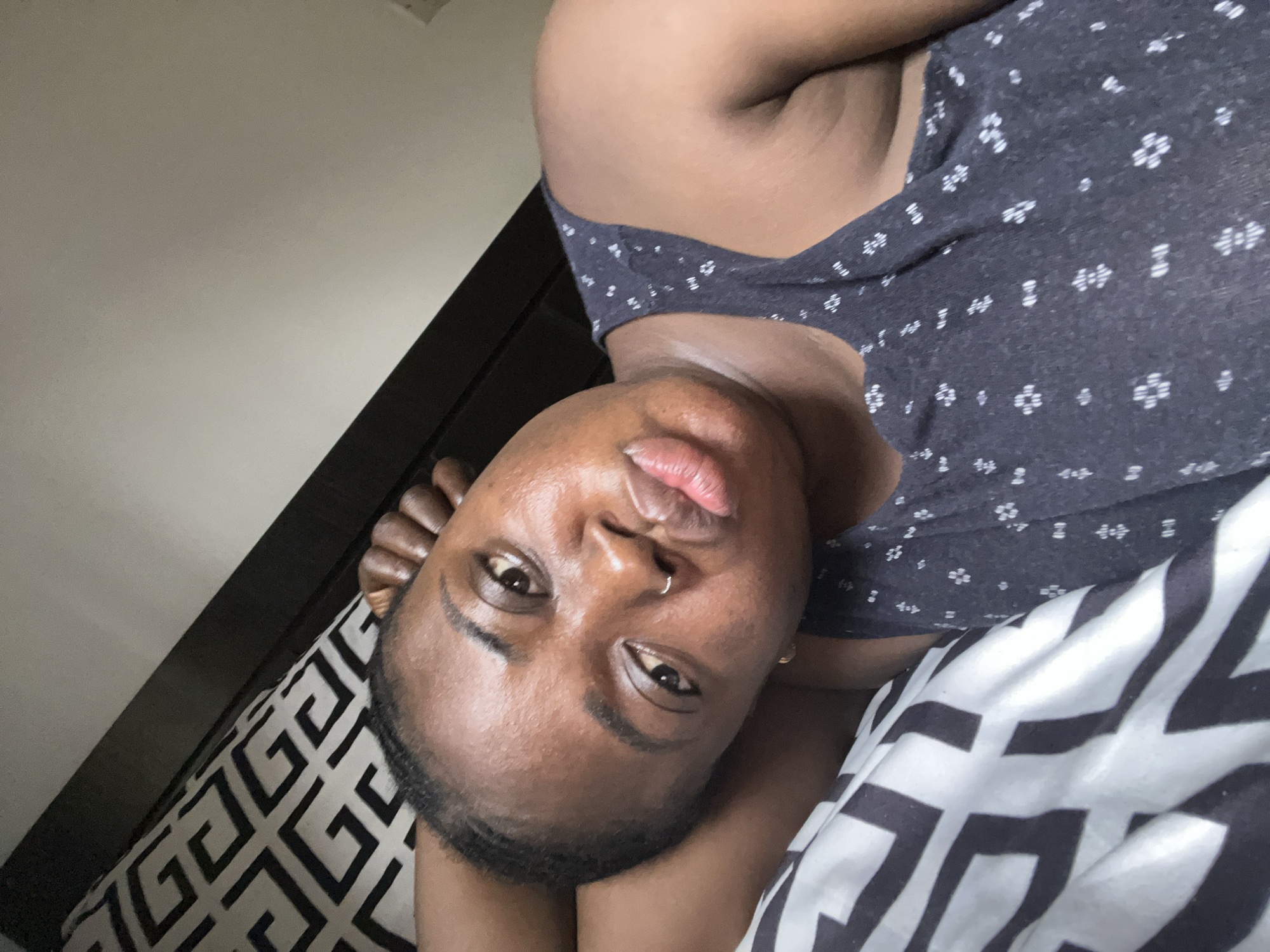 A person is lying down on a bed with patterned sheets. The person has dark skin and is wearing a dark tank top with small white floral patterns. The background includes a plain wall and a dark-colored headboard. The individual has a nose ring and appears to be looking at the camera. The location cannot be determined from the image but it is presumed to be in Abuja, Nigeria.