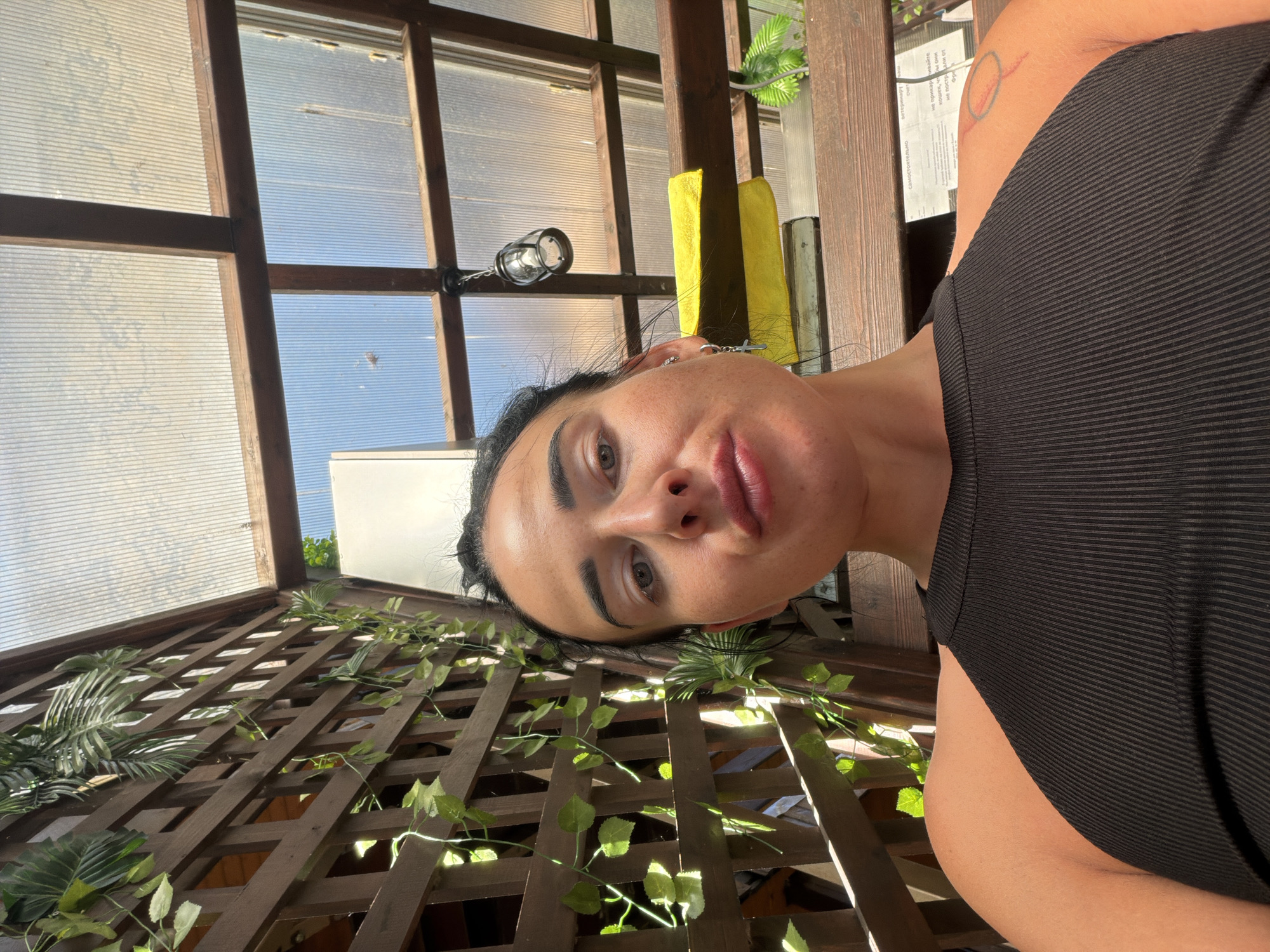 The image shows a close-up, selfie-style shot of a woman in an outdoor, covered seating area. She is wearing a black ribbed tank top. Behind her, a wooden lattice fence with artificial greenery is visible, and above that, a translucent, ribbed roof allows sunlight and a blue sky to filter through. A hanging lamp is visible in the upper right portion of the image, and a yellow towel is draped over a wooden beam. The woman has dark hair pulled back, is wearing a cross-shaped earring, and has a tattoo on her right shoulder. The scene appears to be a casual outdoor setting, possibly a cafe or a patio. The location is not specified in the image.